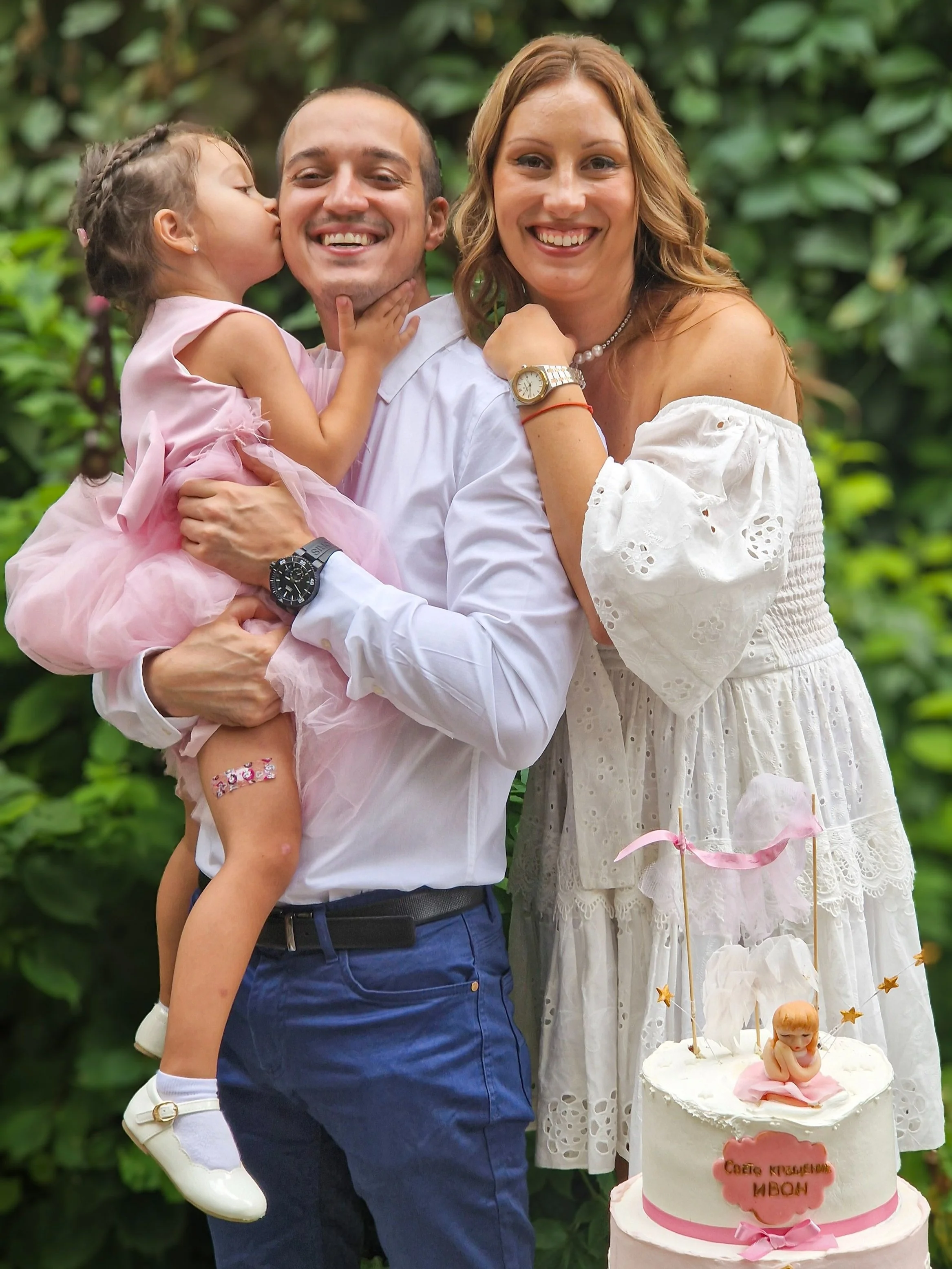 A family celebrating a special occasion outdoors, with a young girl kissing a smiling man on the cheek. The woman beside them smiles at the camera, and a decorated cake is seen in the foreground.