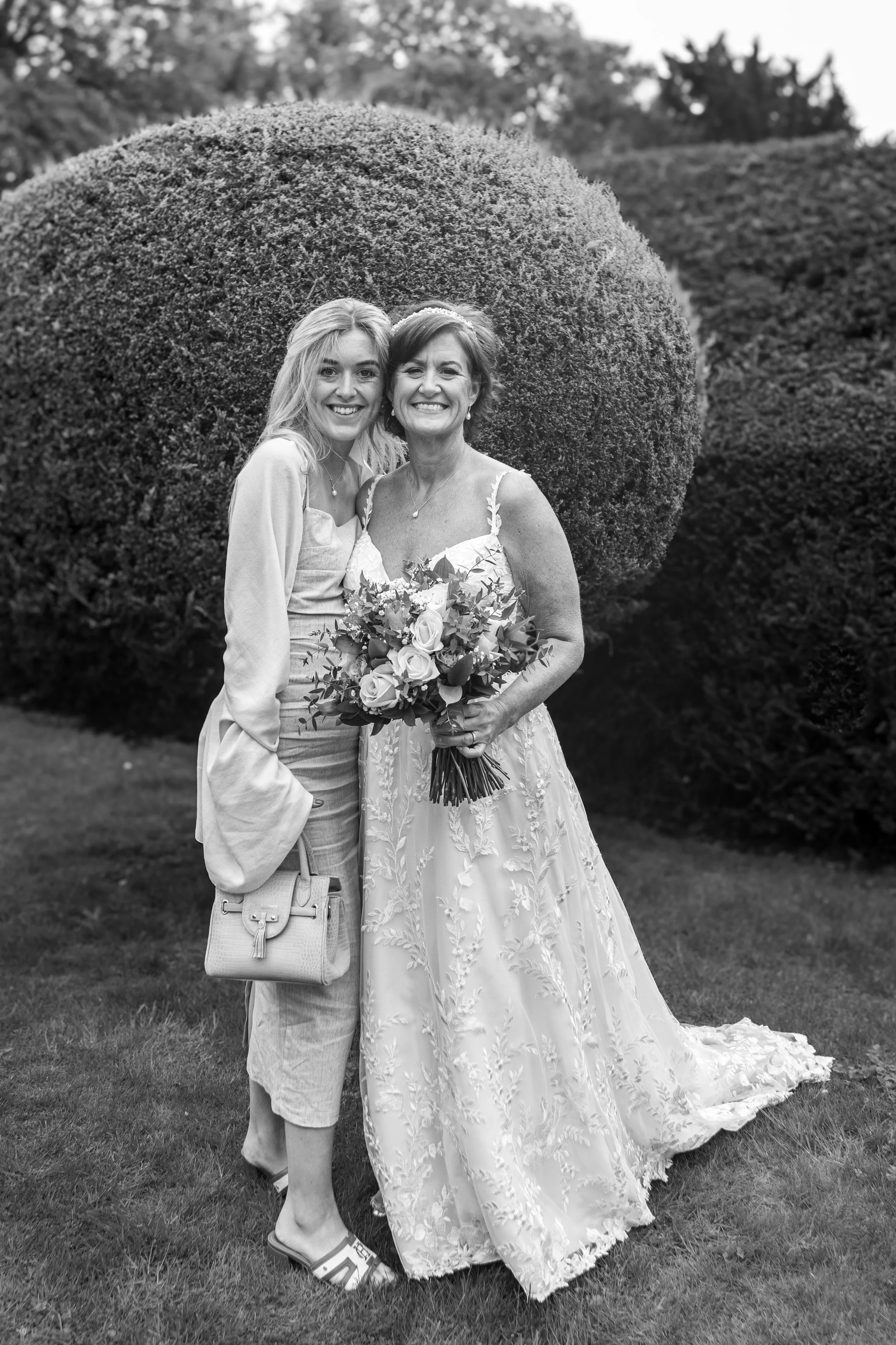 A woman in a wedding dress holding a bouquet standing next to a younger woman, both smiling, outdoors with trimmed bushes in the background.
