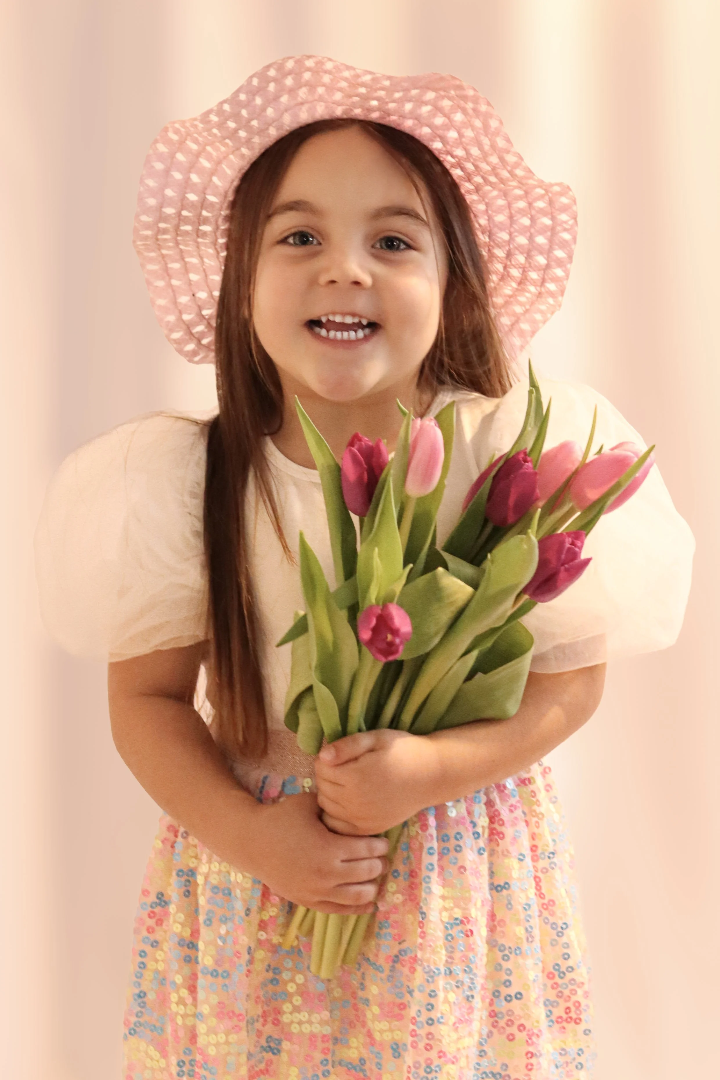 A young girl wearing a pink sunhat and a pastel dress, holding a bouquet of pink tulips, smiling at the camera.
