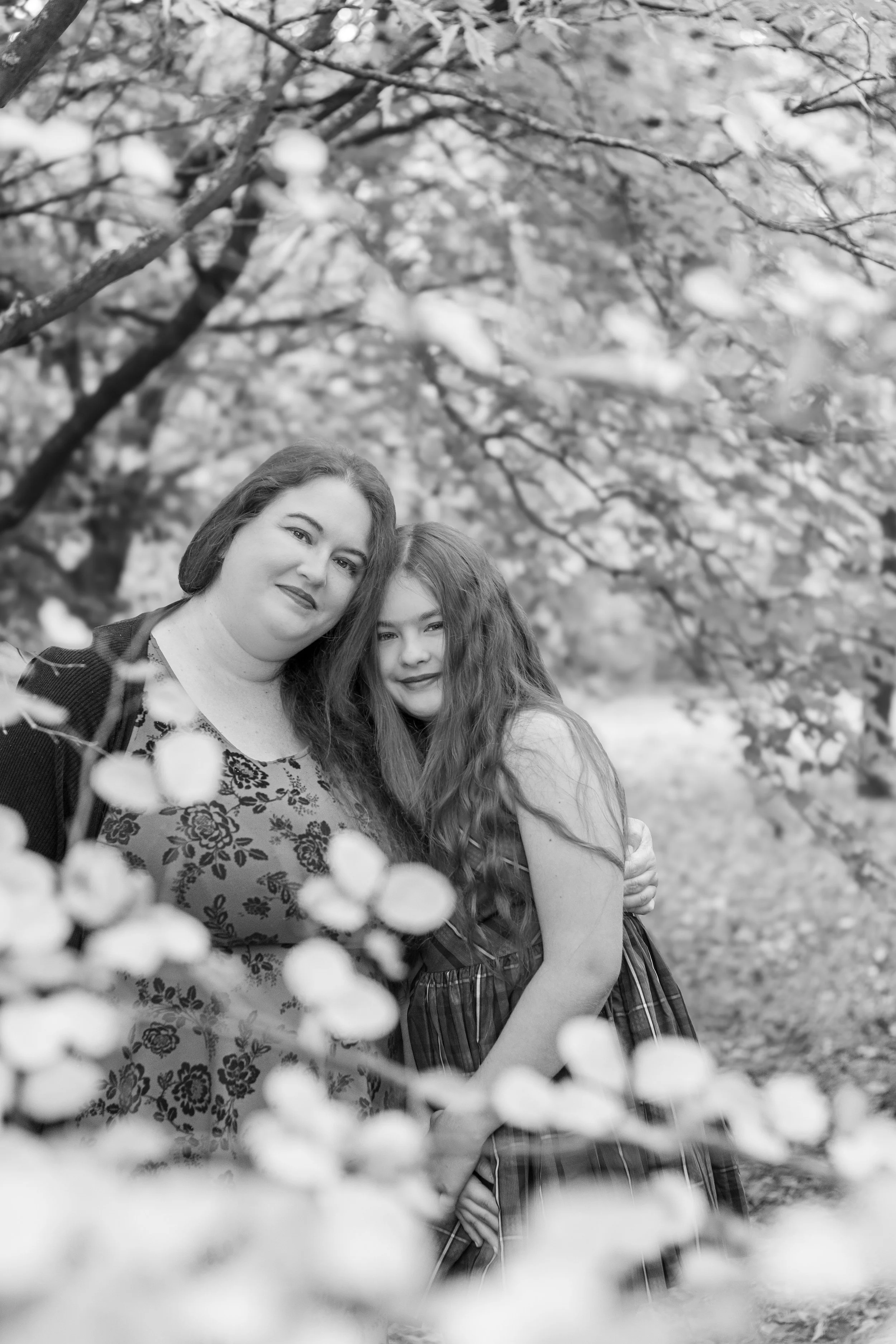 Black and white photo of a woman and a young girl embracing outdoors under tree branches with leaves.