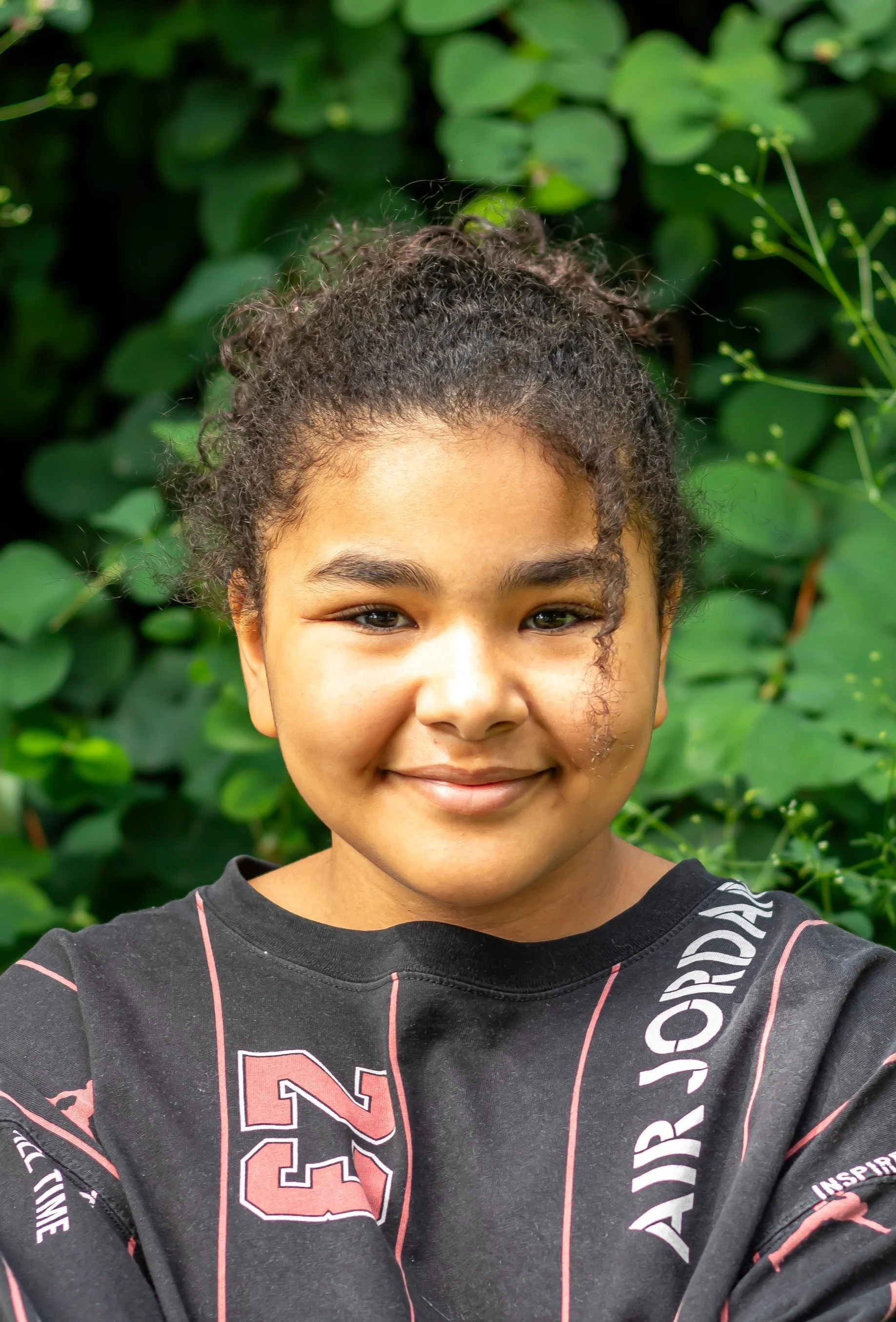 Close-up of a smiling young girl with curly hair and a black shirt with pink and white lettering, standing in front of green foliage.