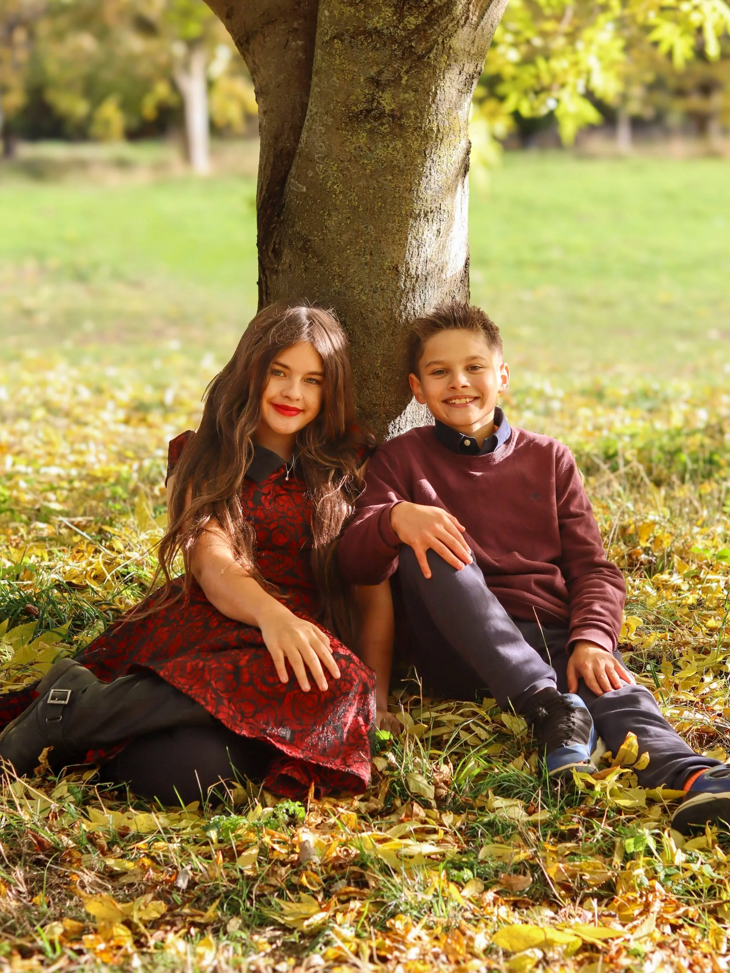 A boy and girl sitting and leaning against a tree on a bed of fallen leaves in a park.