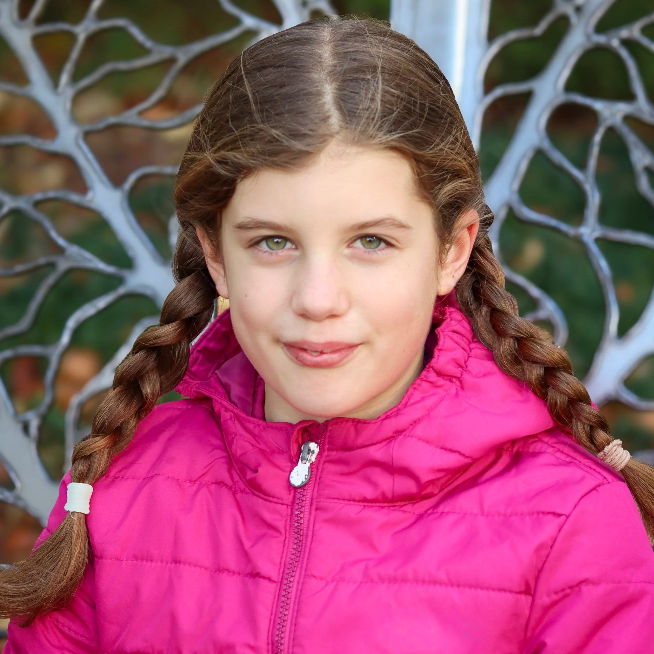 A young girl with braided hair and green eyes, wearing a pink jacket, sitting outdoors on a decorative metal chair.