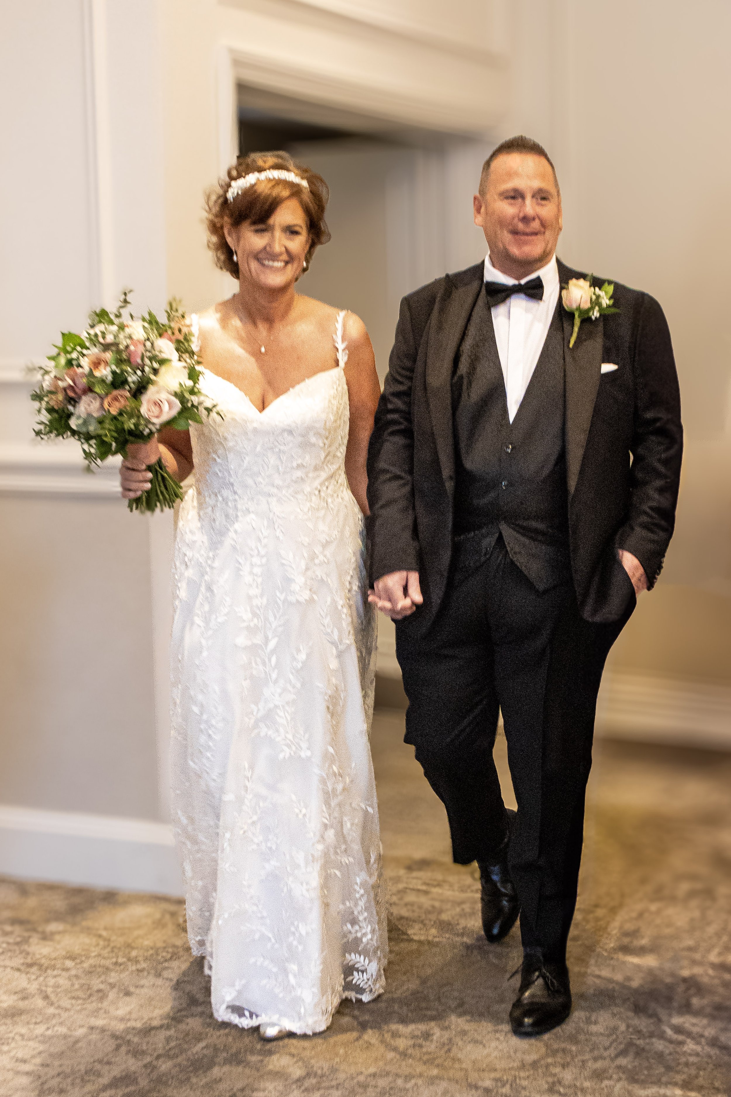 A bride in a white lace wedding gown holding a bouquet, walking with a groom in a black tuxedo. They are indoors, smiling, with the groom holding the bride's hand.