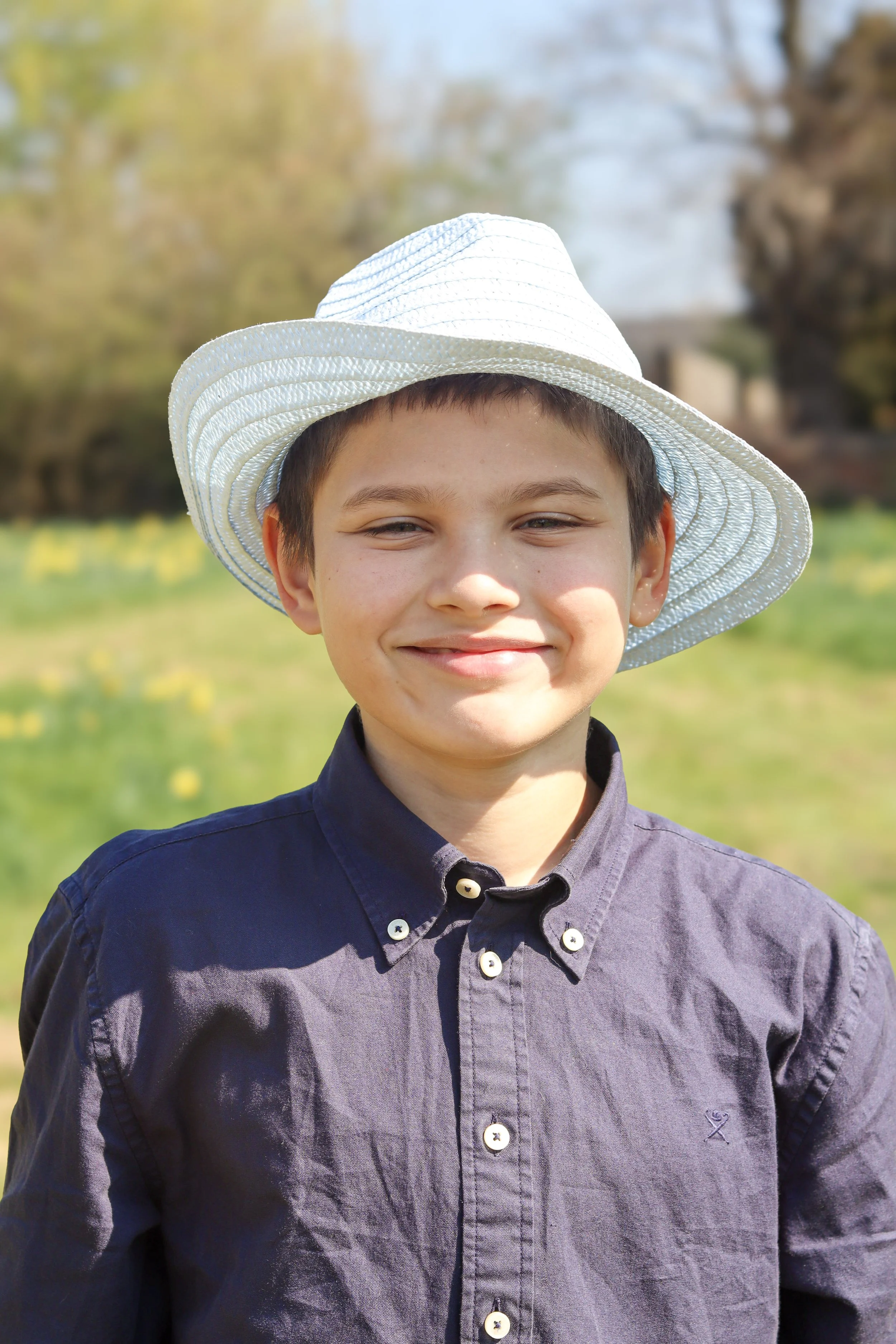 A young boy smiling outdoors, wearing a white sun hat and a button-up shirt on a sunny day.