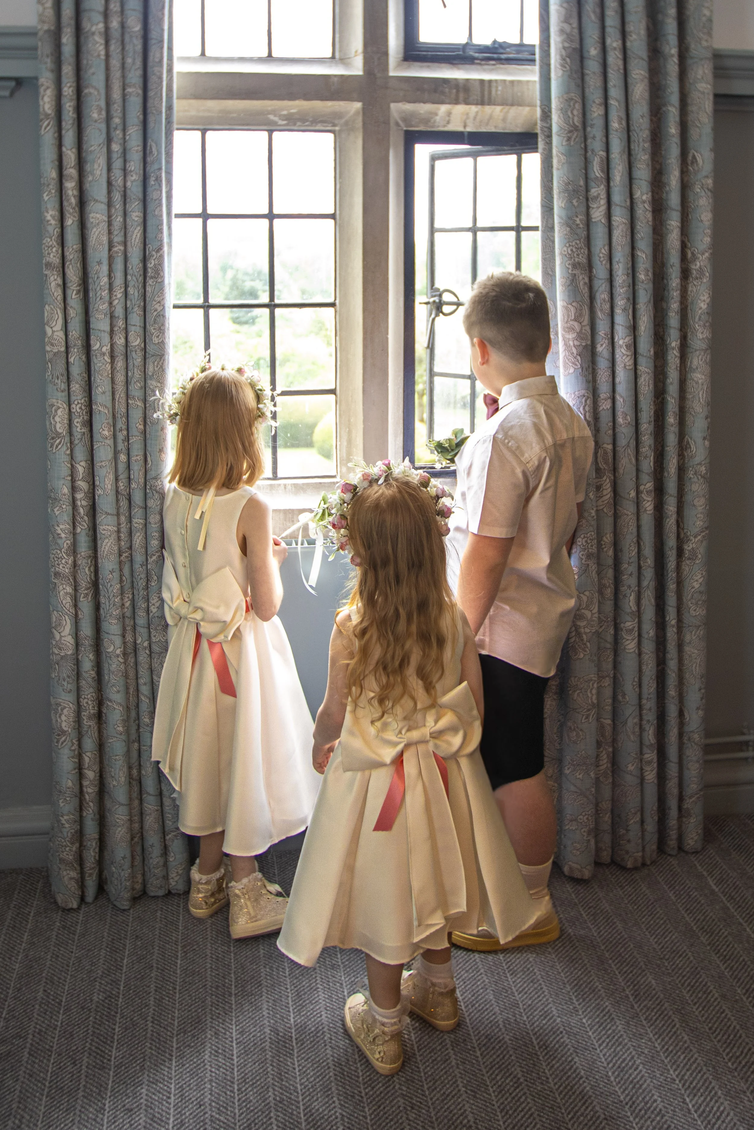 Three children dressed in formal attire, with two girls wearing wedding dresses and a boy in a shirt and shorts, looking out the window and holding flower crowns.