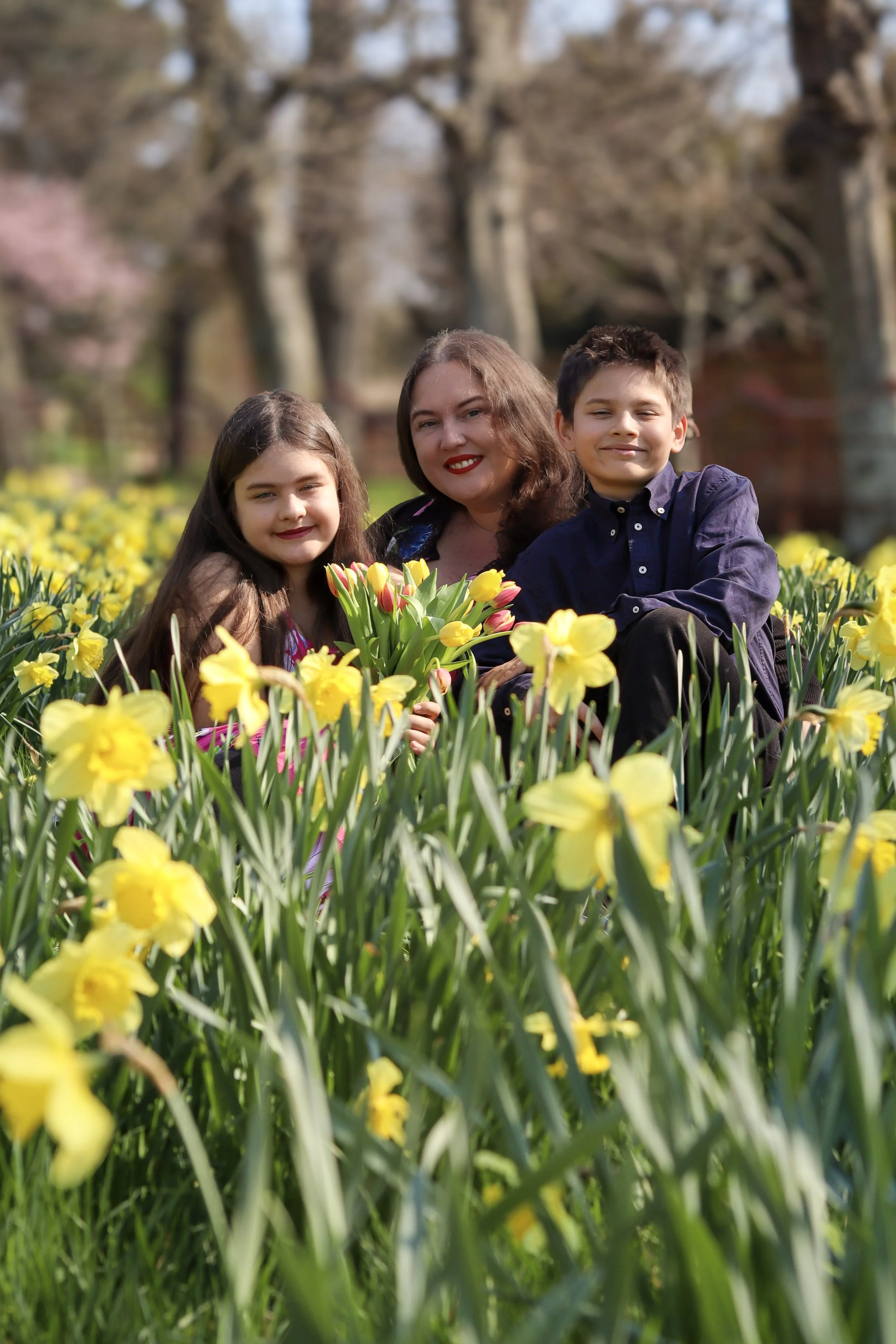 A woman and two children sitting in a field of yellow and pink tulips, smiling at the camera, with trees in the background.
