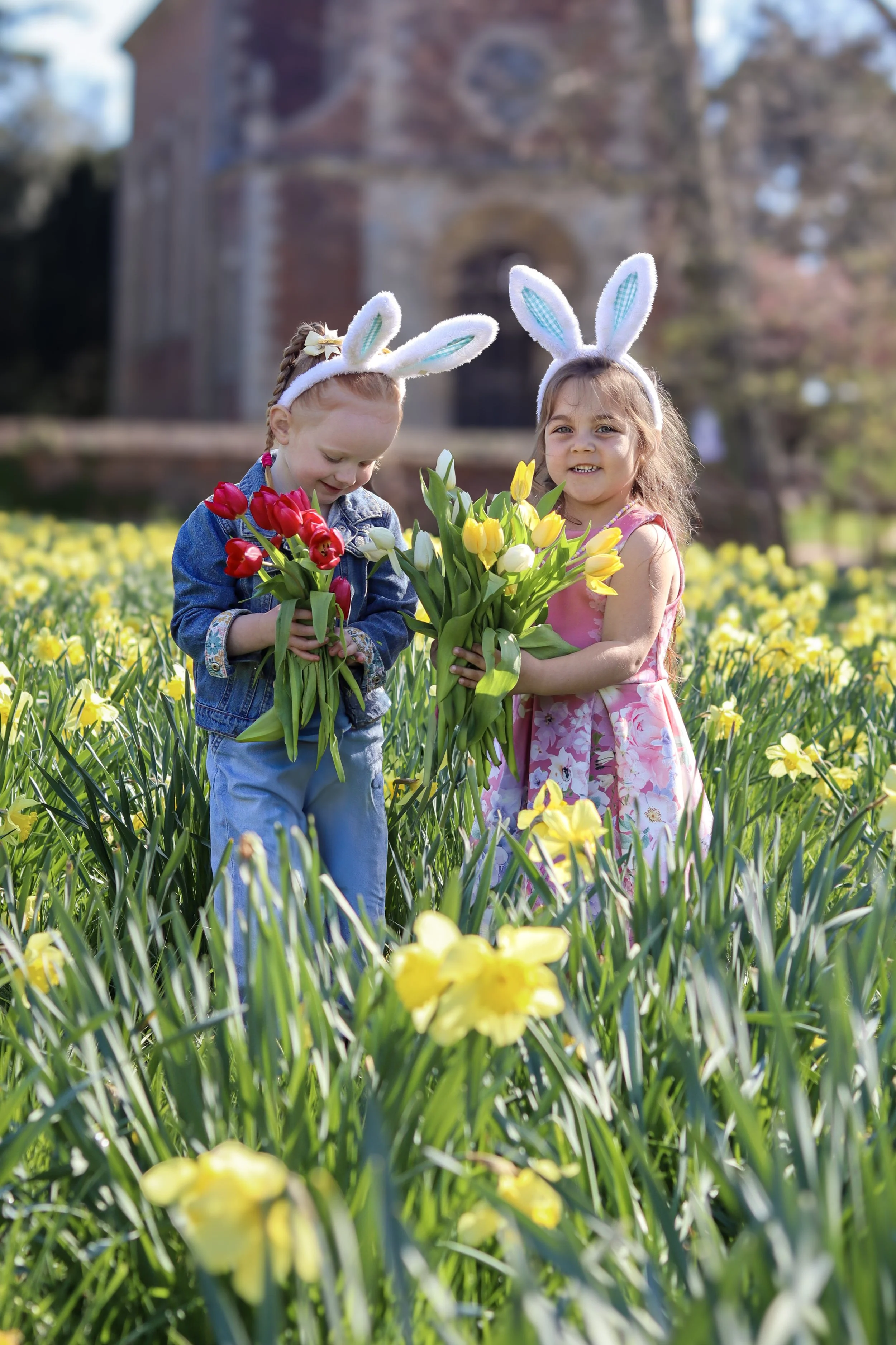 Two young girls wearing bunny ear headbands standing in a field of yellow and white flowers, holding bouquets of tulips, with a historic building in the background.