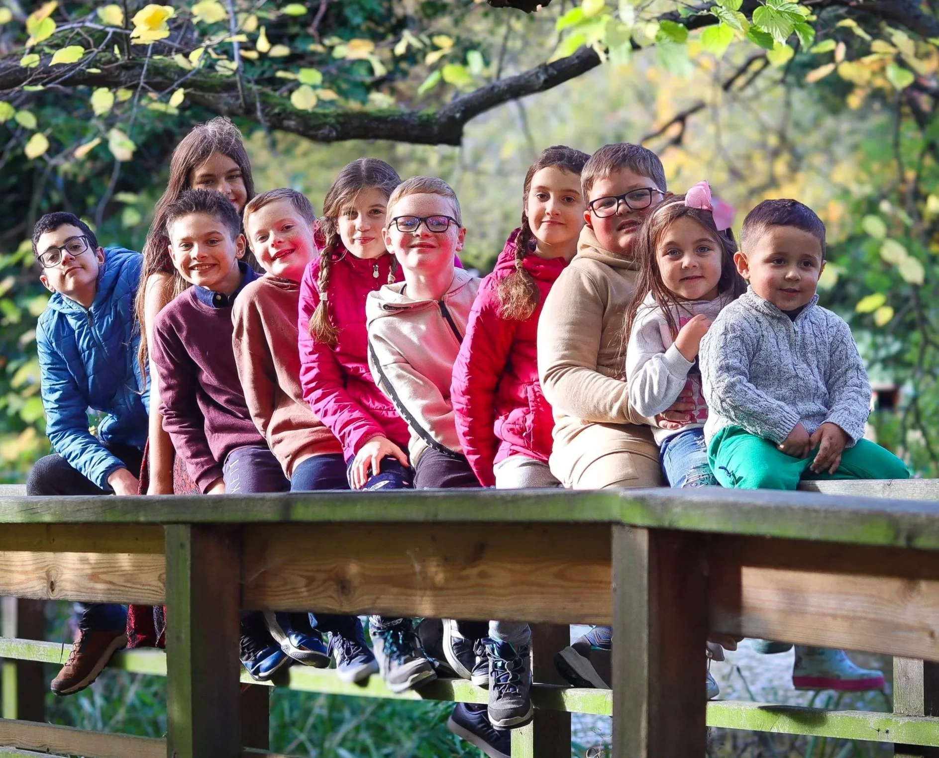 A group of children sitting in a row outdoors on a wooden platform, surrounded by green trees with yellow leaves in the background.