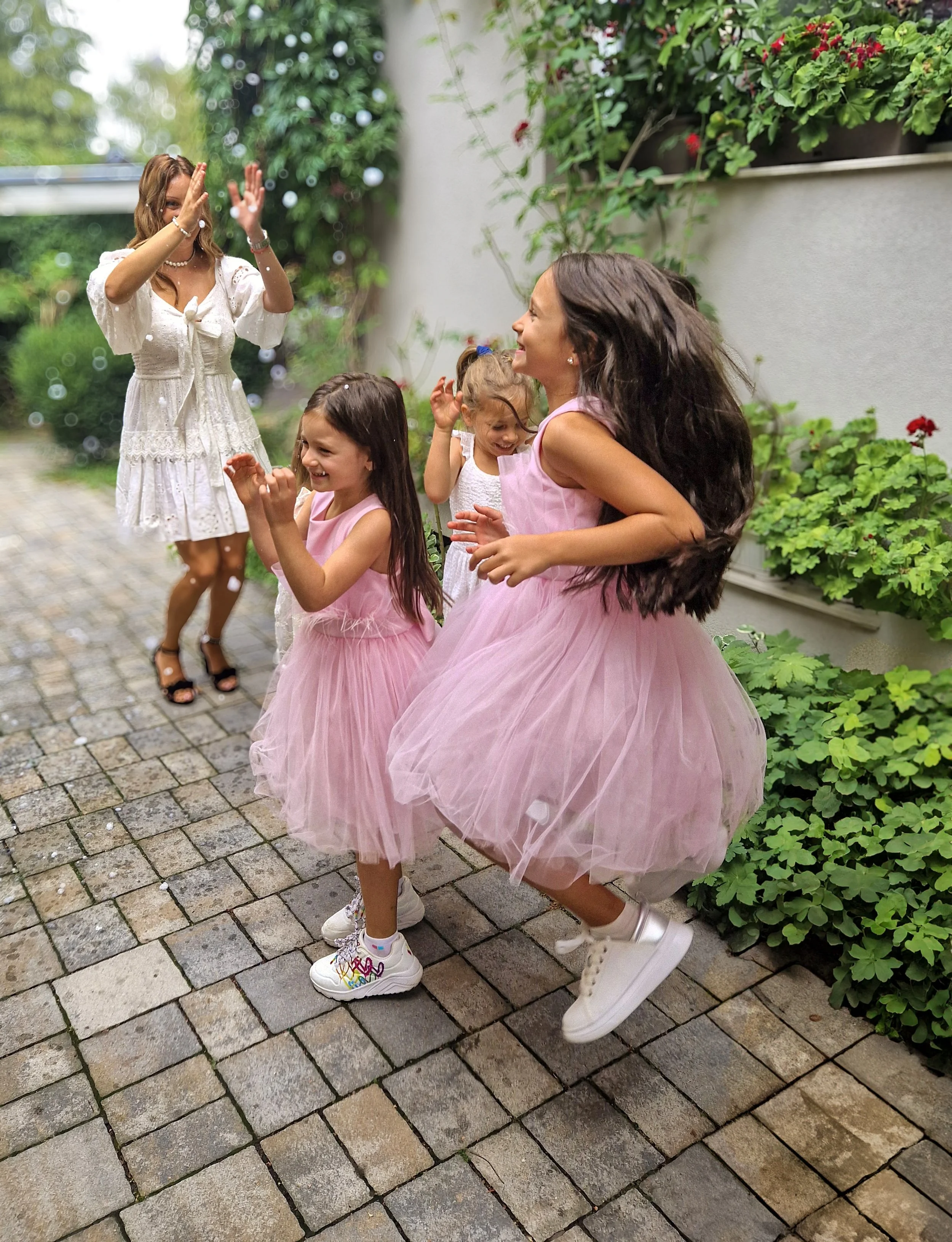Four children and an adult girl dancing and having fun outdoors on a brick pathway, surrounded by greenery and flowering plants.