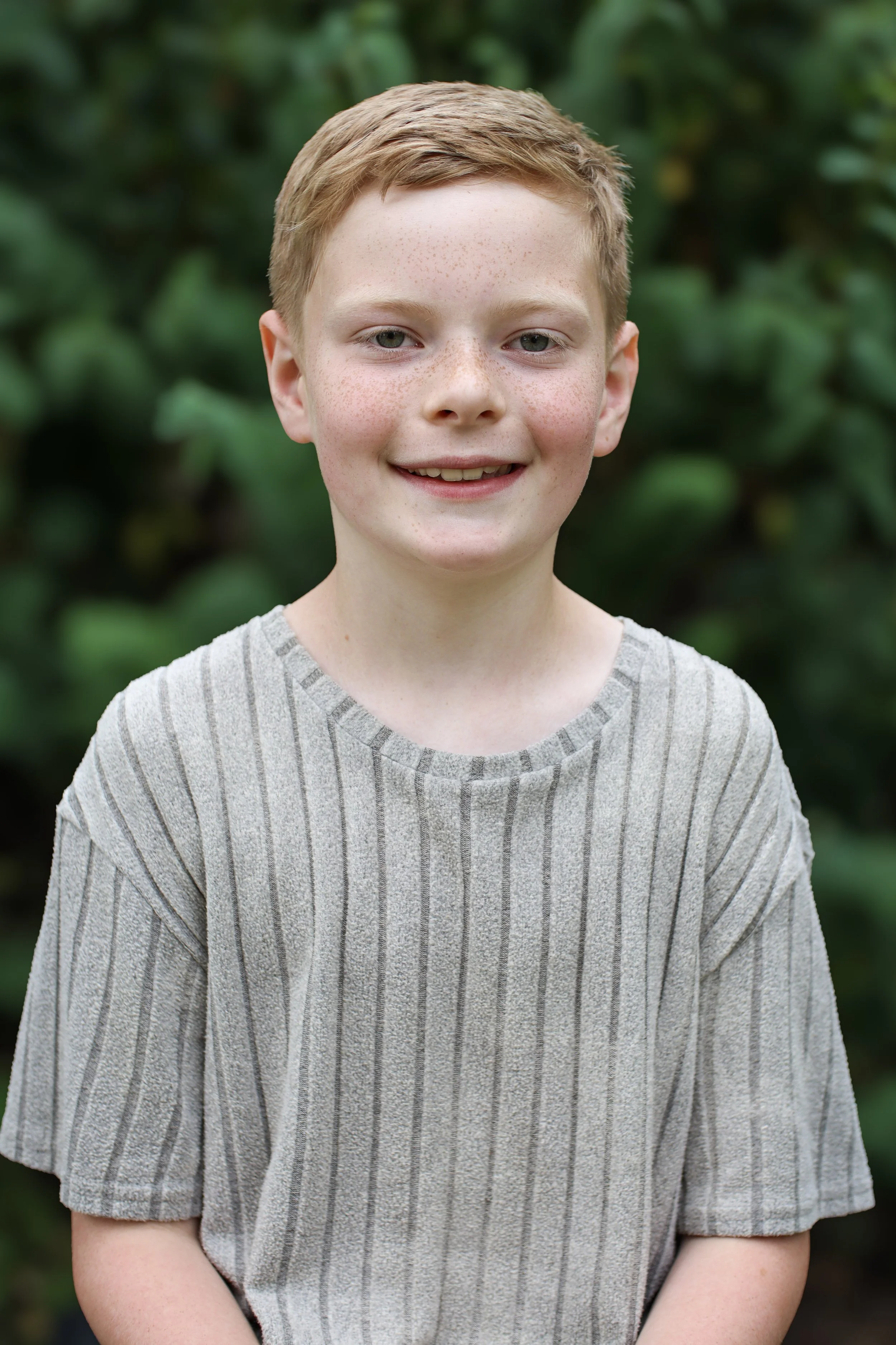 A young boy with short red hair, freckles, and a light gray and white striped t-shirt, standing outdoors in front of green foliage, smiling at the camera.