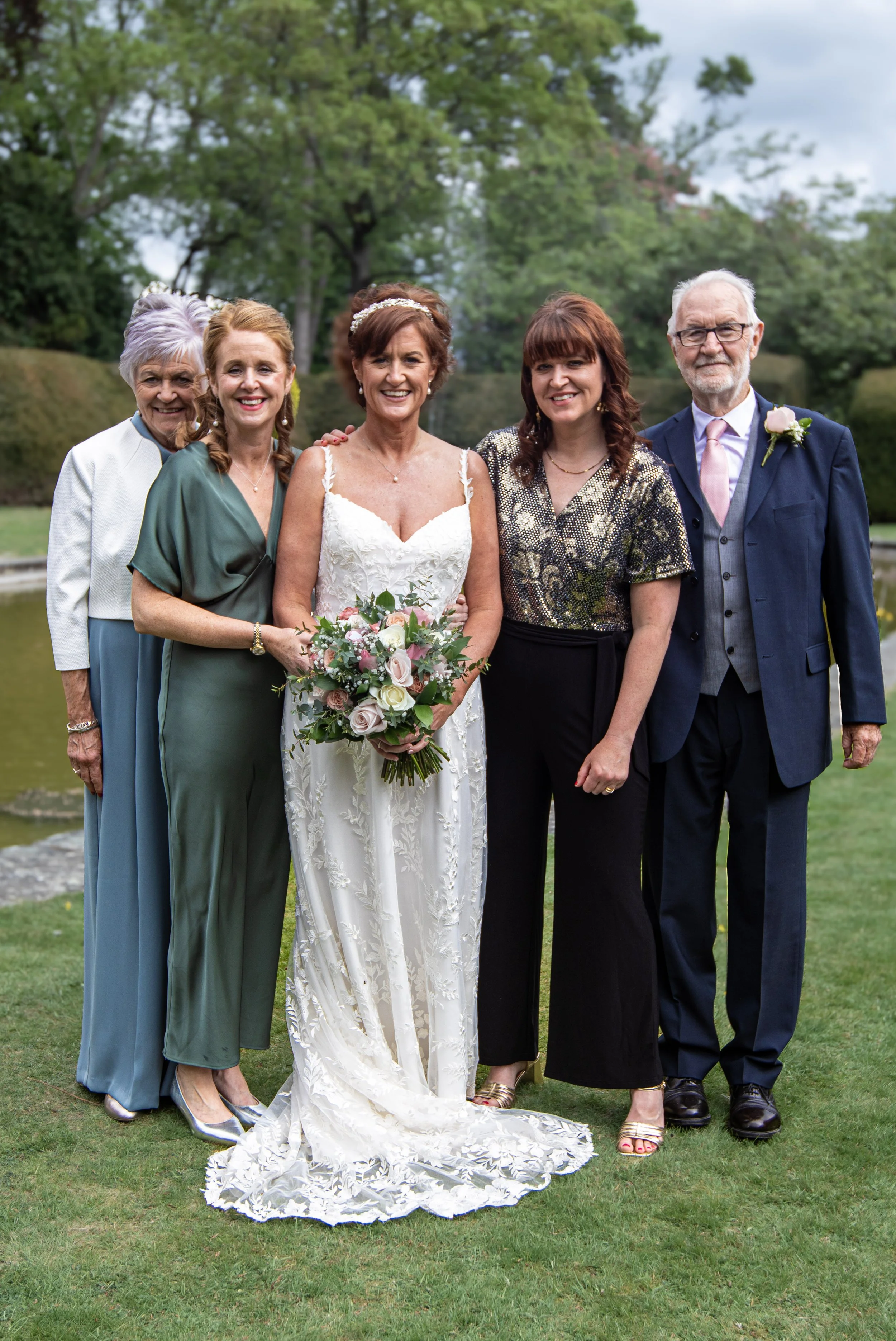 A group of five people, including a bride in a white wedding dress holding a bouquet, standing outdoors on grass with trees in the background.