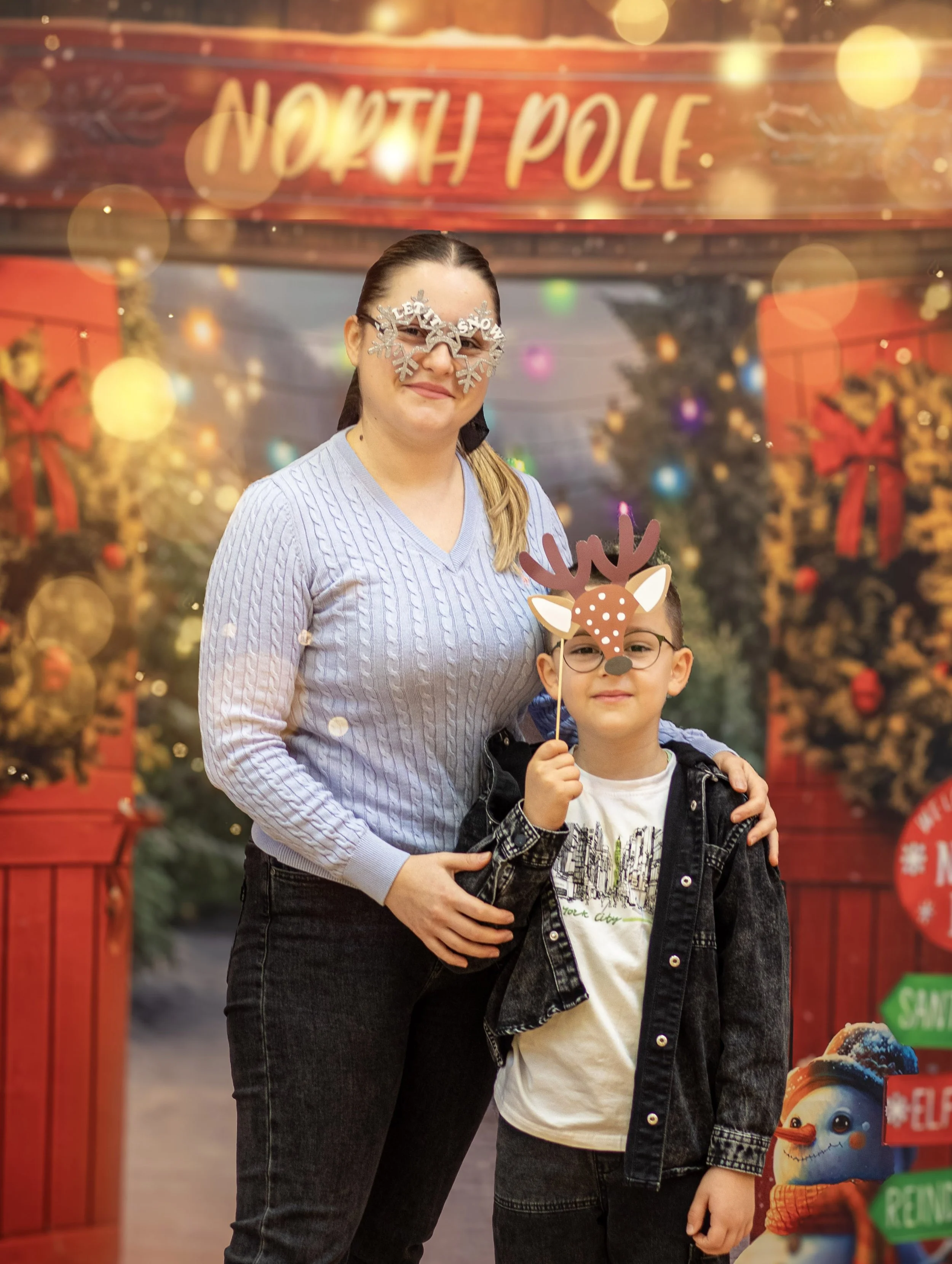 A woman and a young boy at a Christmas-themed photo booth. The woman wears festive snowflake glasses and a light purple sweater, while the boy has reindeer glasses and a Christmas prop, both smiling in front of decorated holiday backdrops.