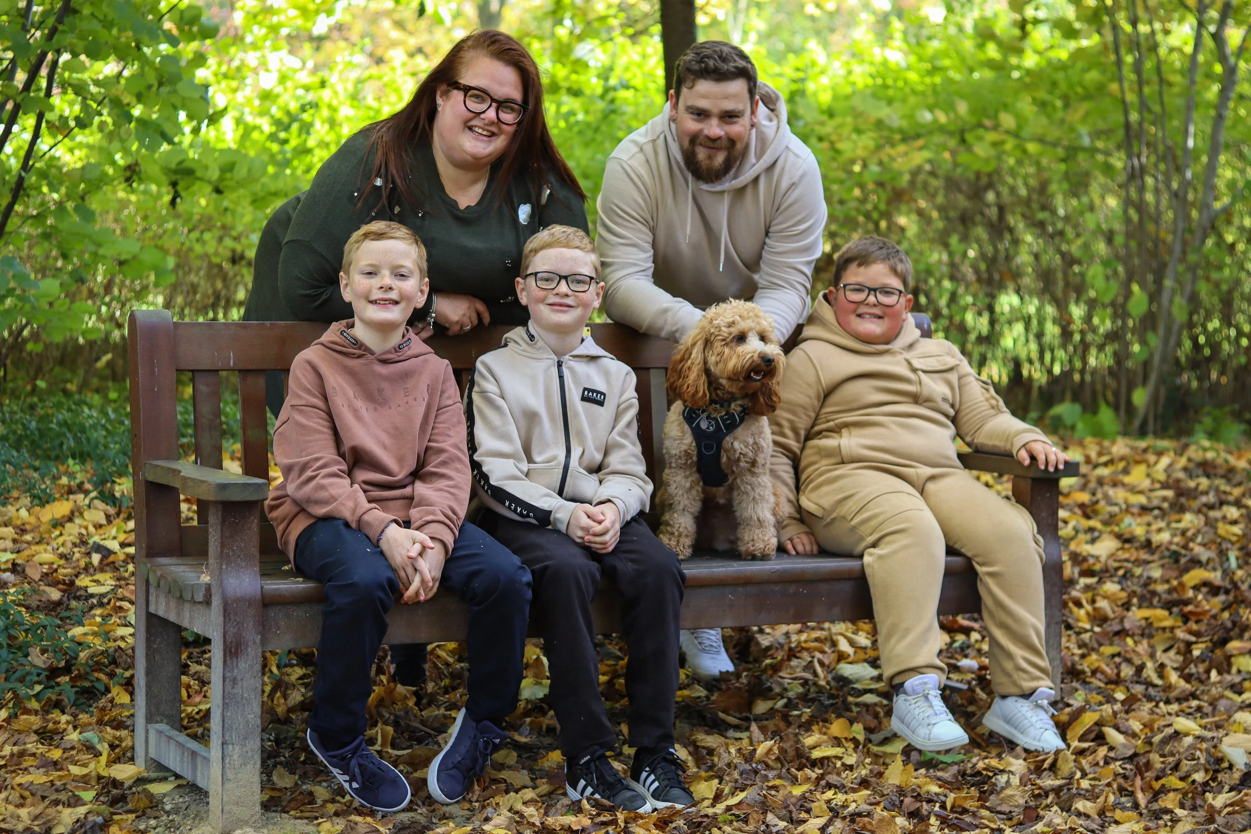 A family of five and their dog sitting on a bench in a park with fallen leaves and green trees in the background.