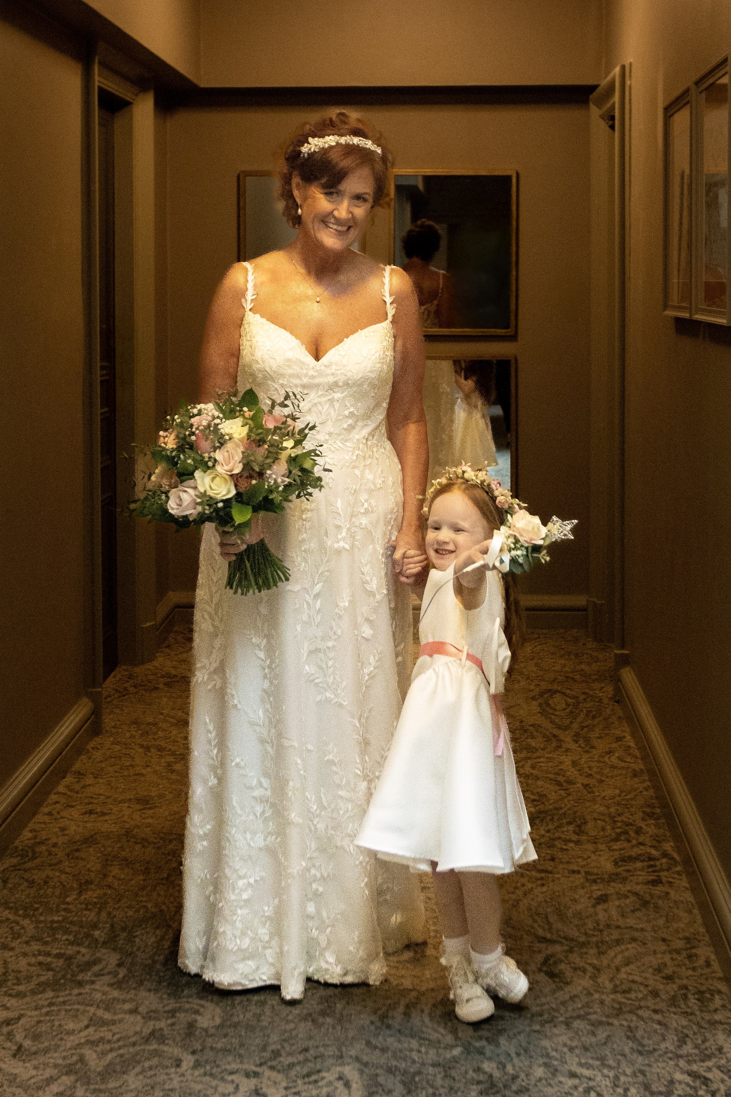 A woman in a white wedding dress holding a bouquet of flowers standing next to a young girl in a white dress and pink sash holding a small bouquet, both smiling and holding hands in a hallway.
