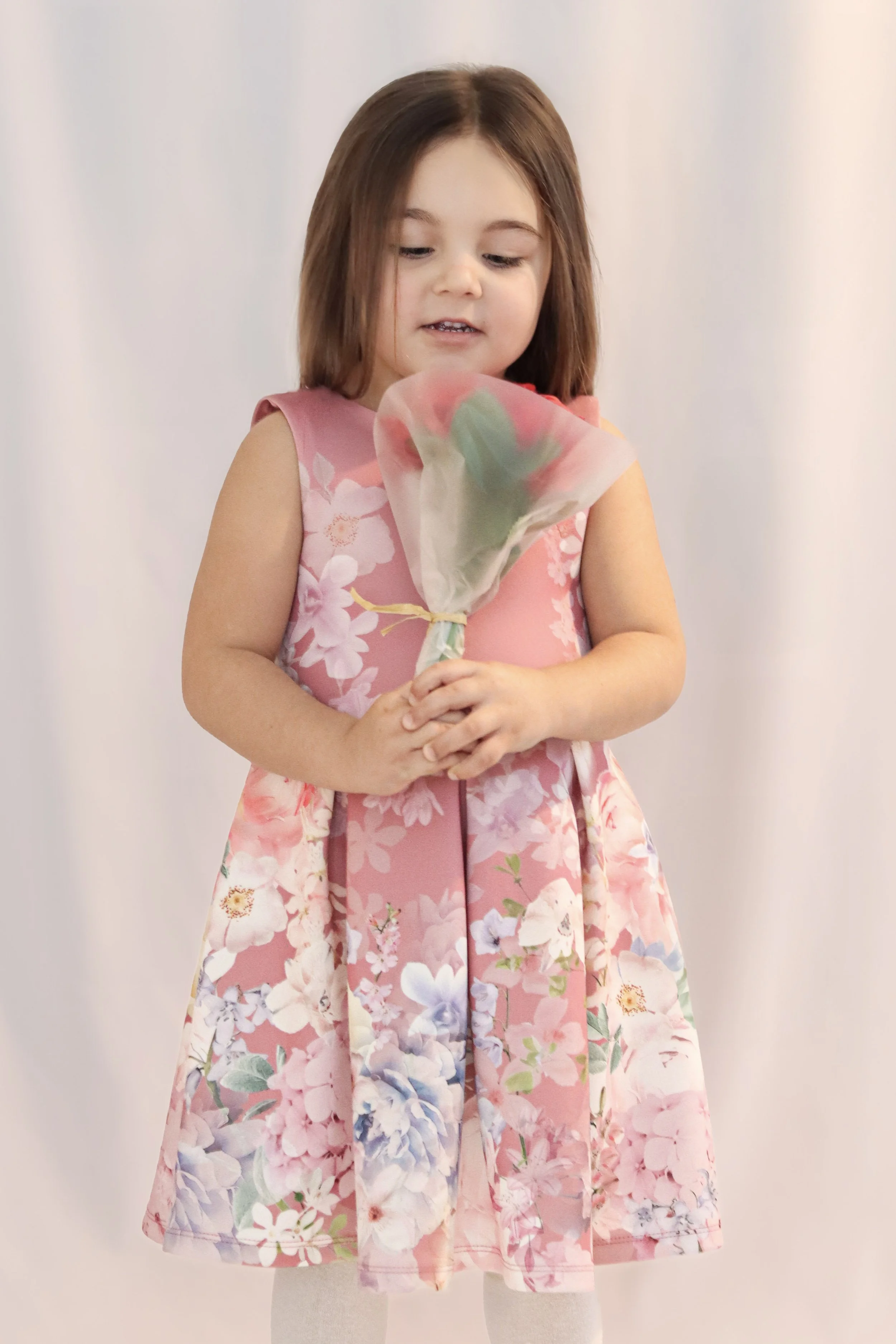 A young girl with brown hair wearing a pink floral dress, holding a wrapped bouquet of flowers, standing in front of a plain white backdrop.