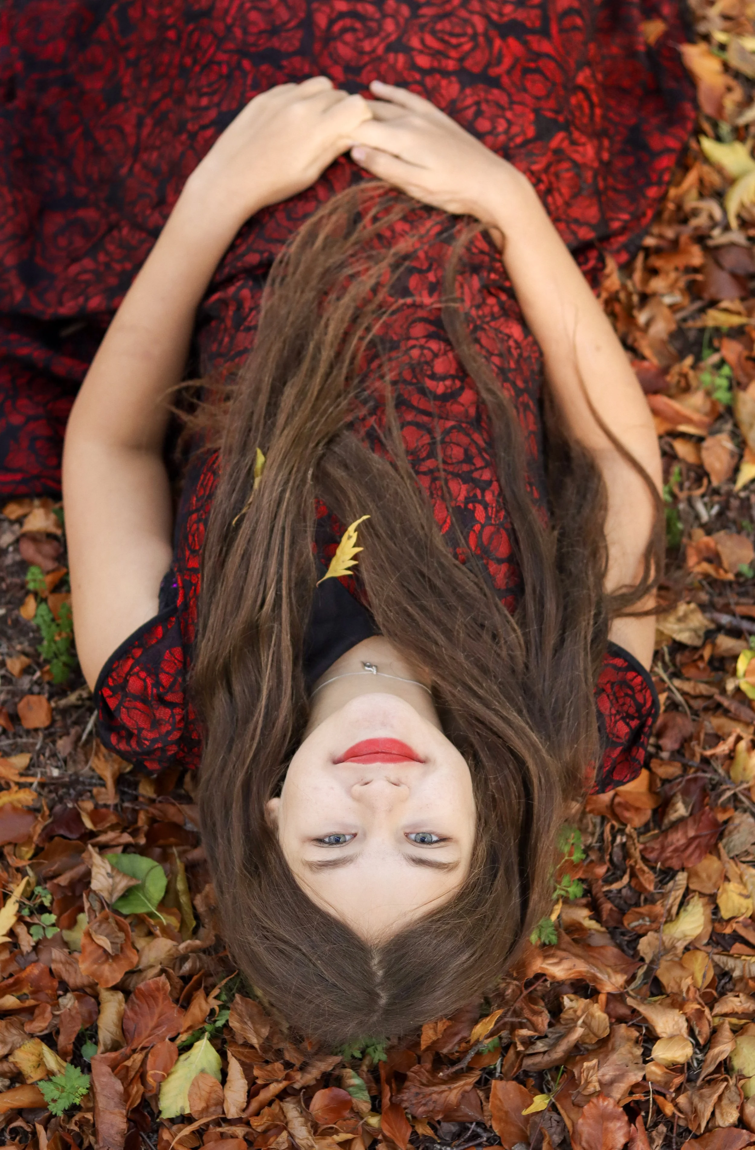A woman with long brown hair and red lipstick lying on the ground covered with autumn leaves, wearing a black and red lace dress.
