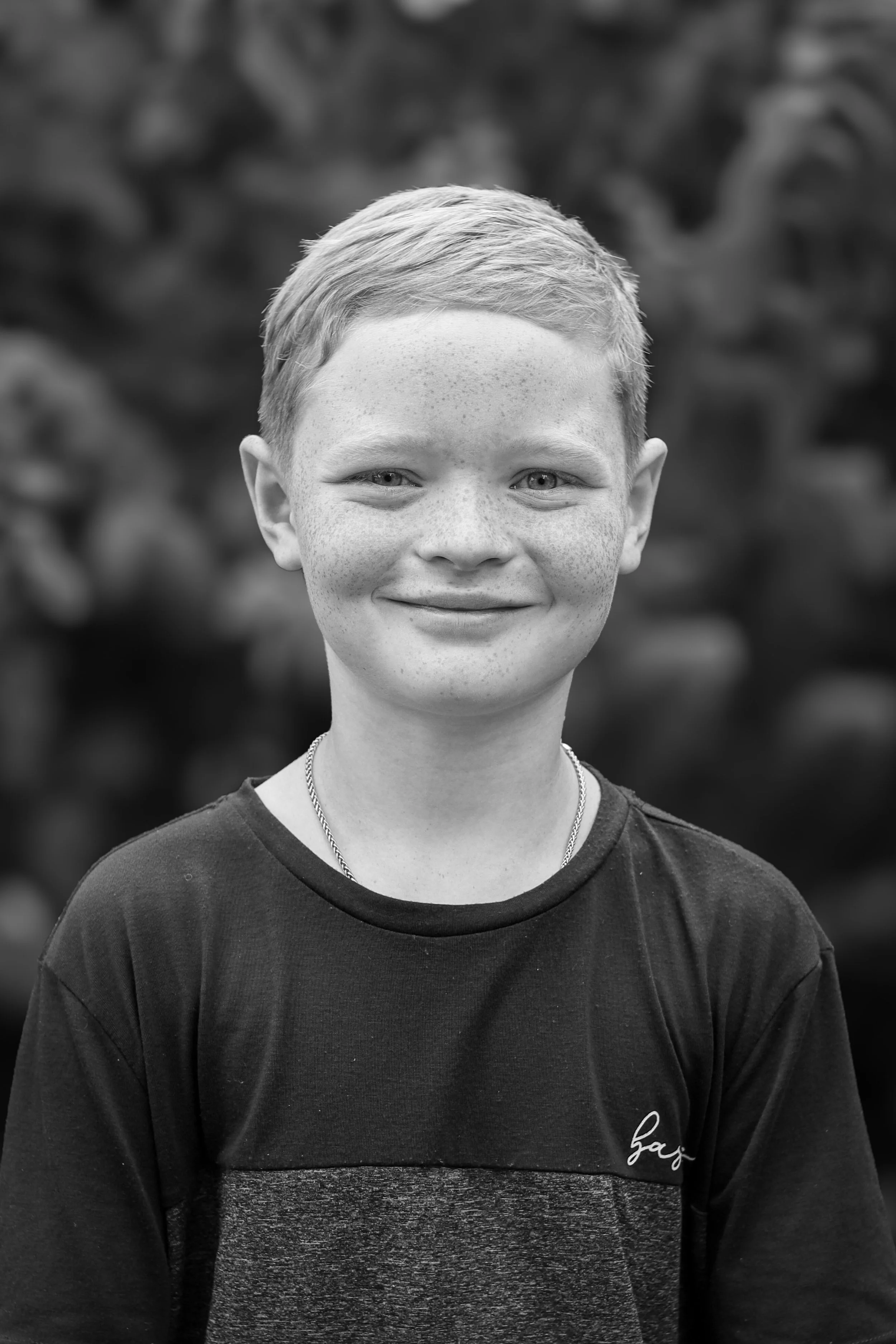 Black and white portrait of a smiling young boy with short hair, wearing a casual shirt and a chain necklace, standing outdoors with a blurred natural background.