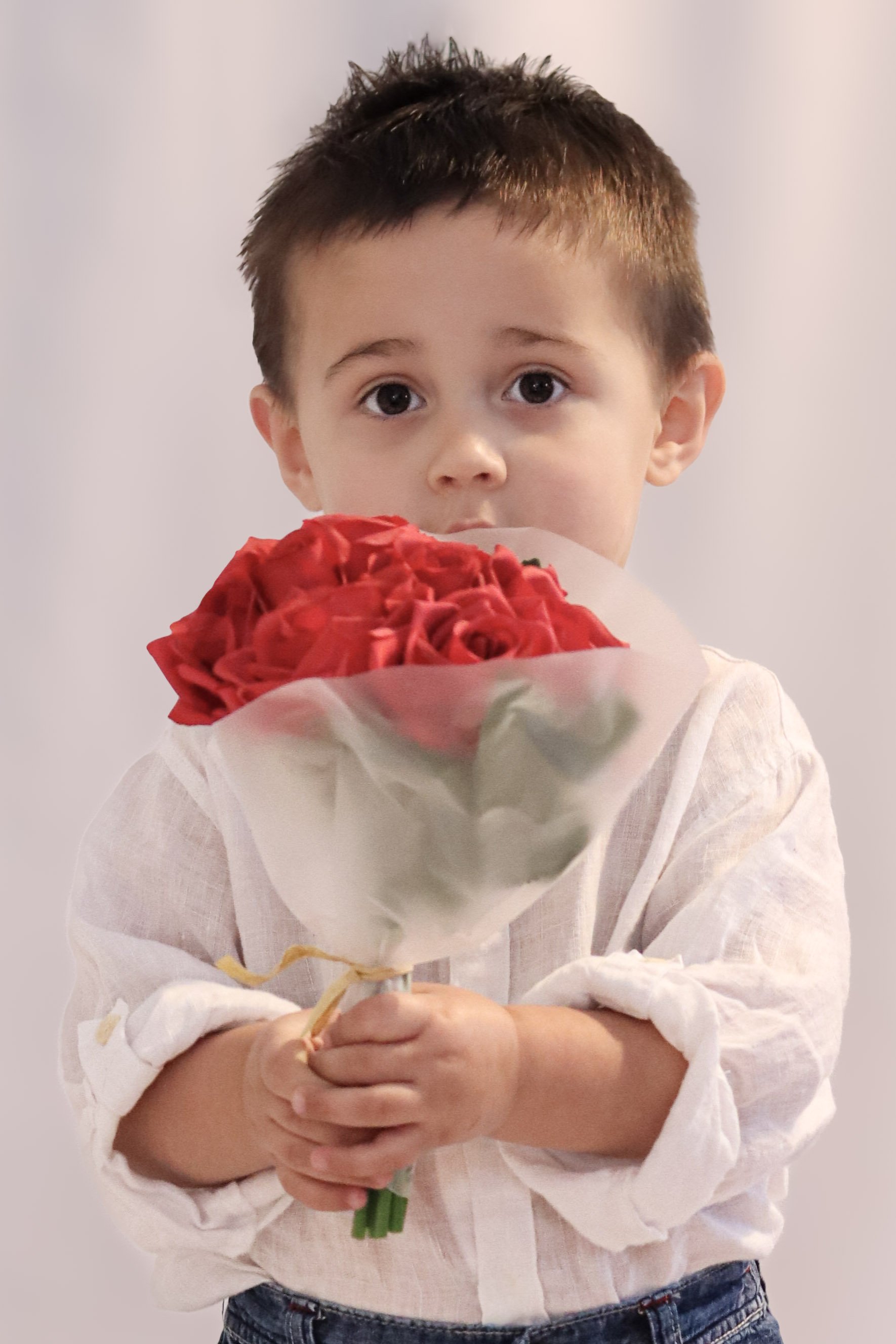 A young boy with short dark hair holding a bouquet of red roses wrapped in paper, looking directly at the camera with a slightly surprised expression, wearing a white shirt and blue jeans.