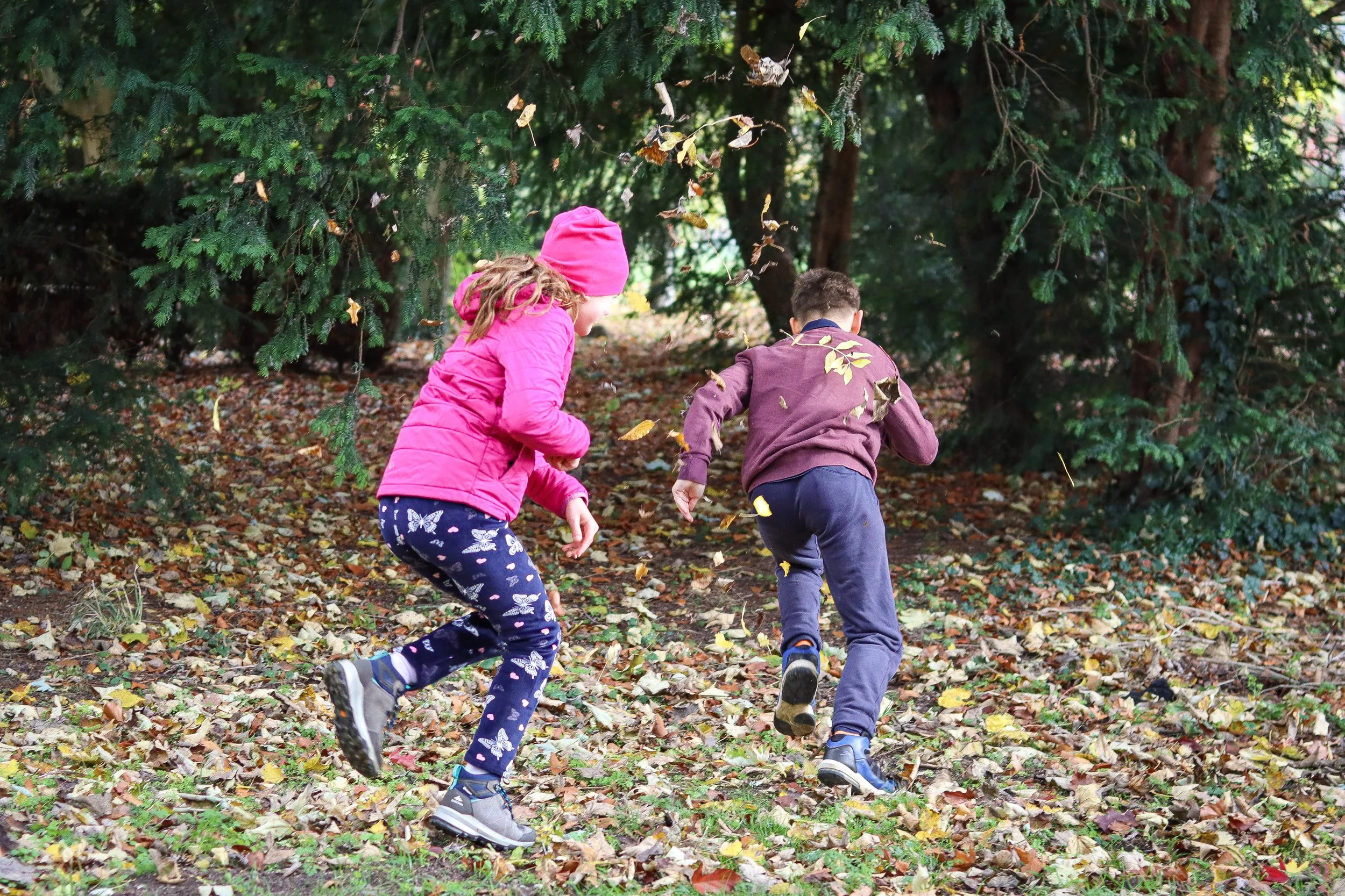 Two children, a girl in a pink jacket and hat, and a boy in a brown jacket, running through a wooded area with fallen leaves.