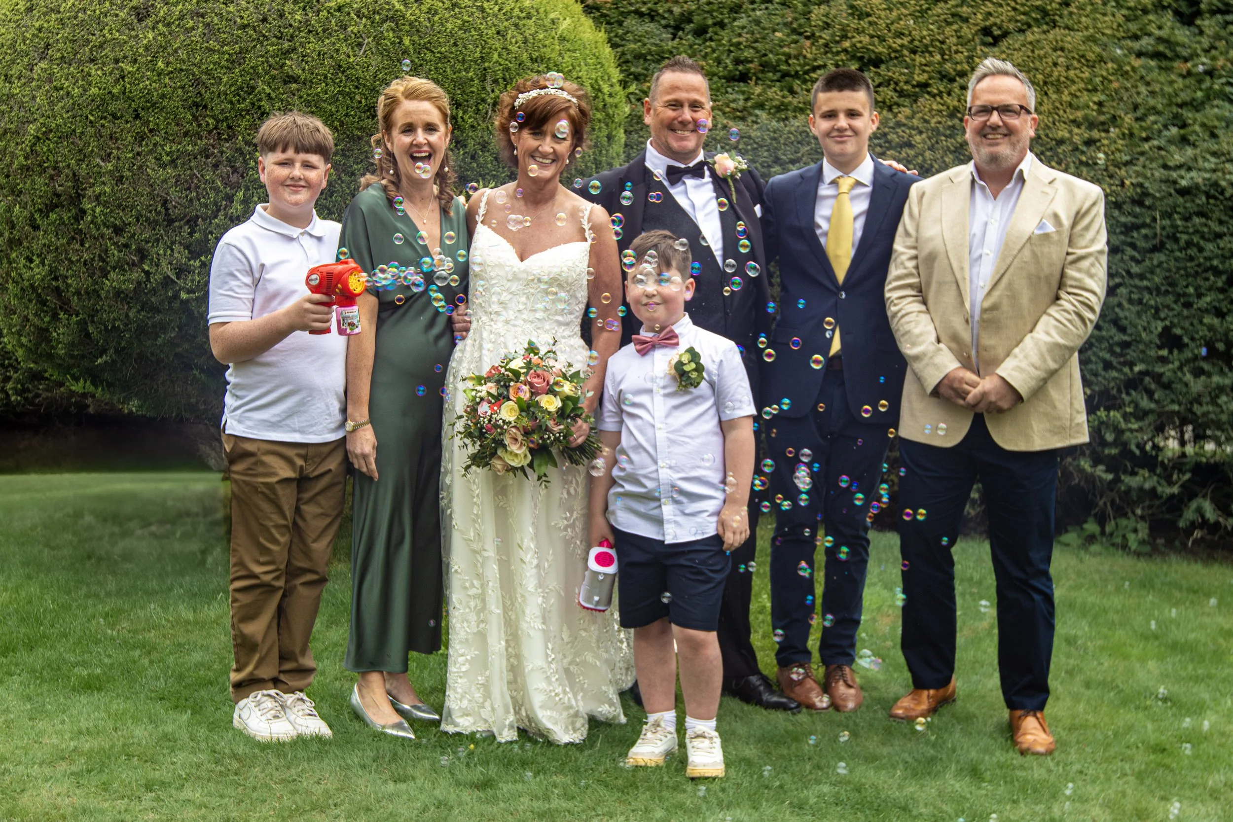Group of people at a wedding, including the bride in a white gown holding a bouquet, the groom in a tuxedo, and five other family members or friends standing outdoors on a grassy area, with bubbles floating around.
