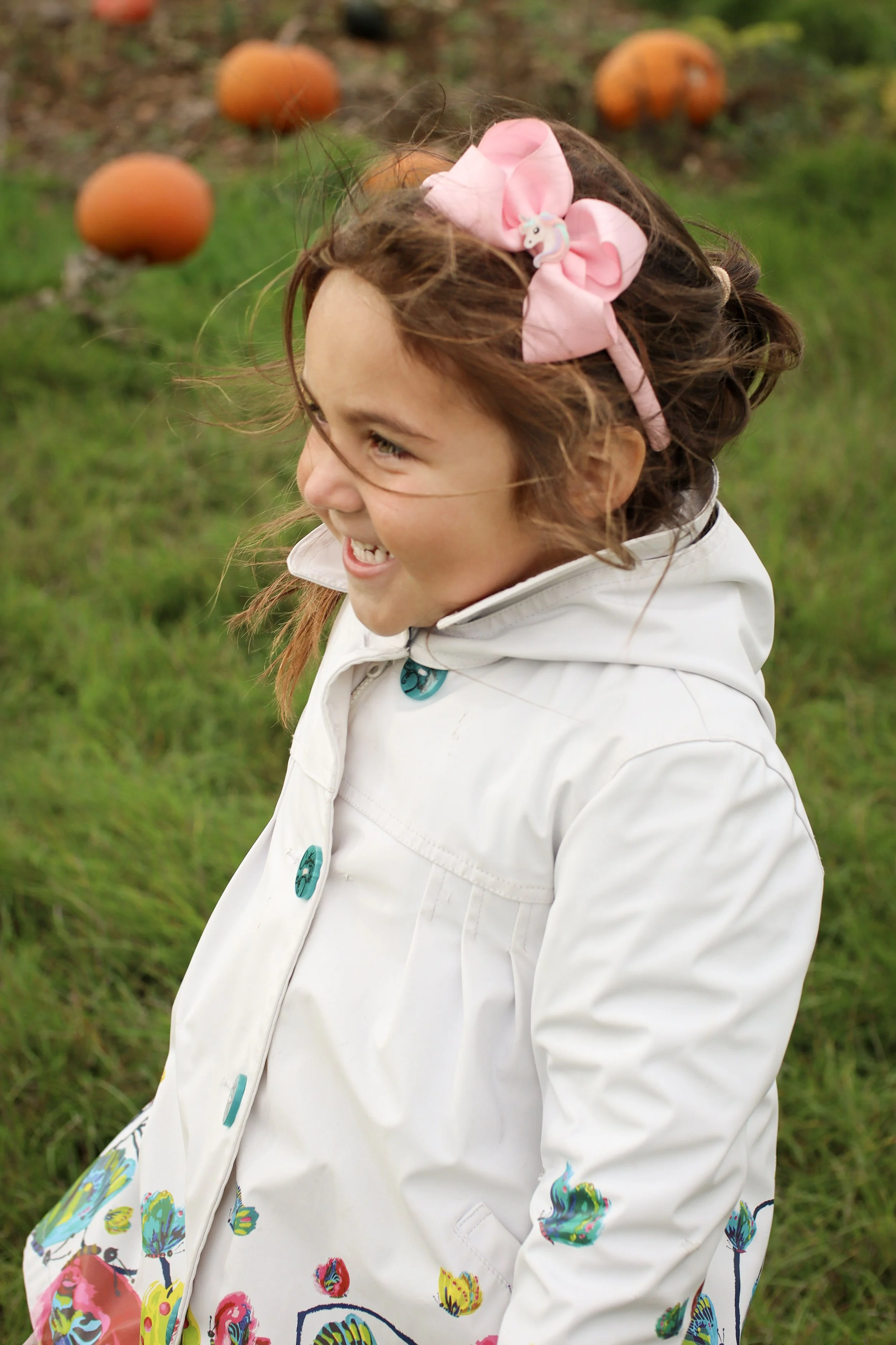 A young girl with dark brown curly hair wearing a white coat with colorful patterns and a pink hair bow, smiling outdoors with pumpkins in the background.