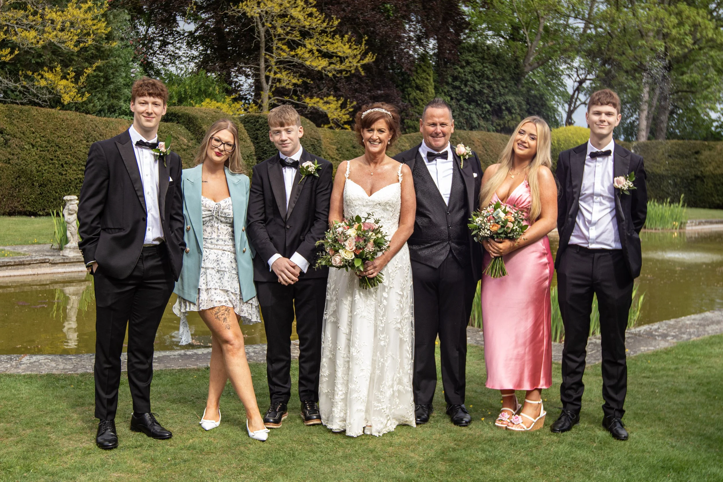 A group of wedding guests, including a bride and groom, standing outdoors in front of a pond and garden.