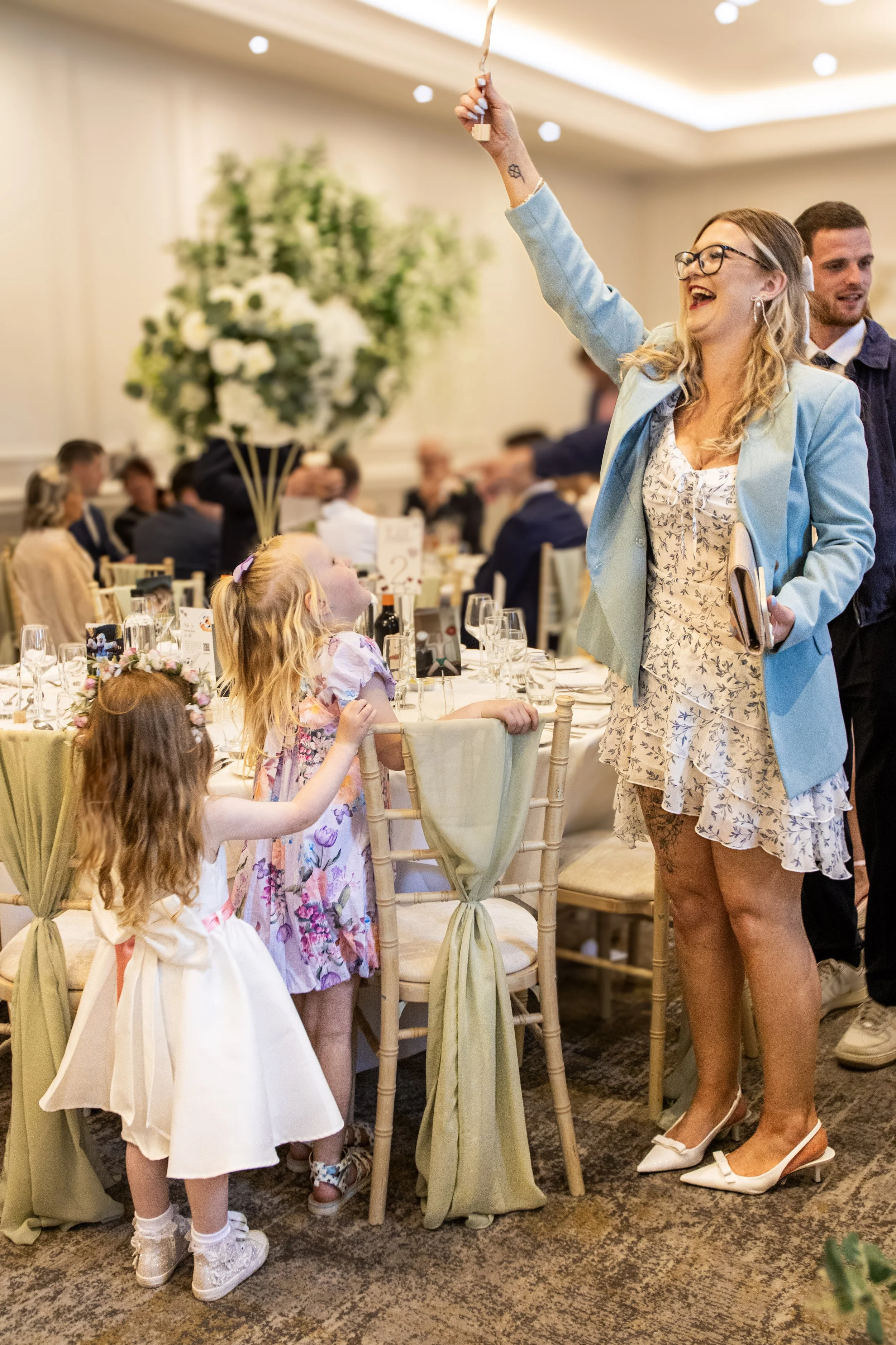 A woman in a light blue blazer and floral dress laughing and raising a glass in a celebratory toast at a banquet table, with two young girls in dresses watching her. The event is held in an elegantly decorated room with a large floral arrangement and many guests in the background.