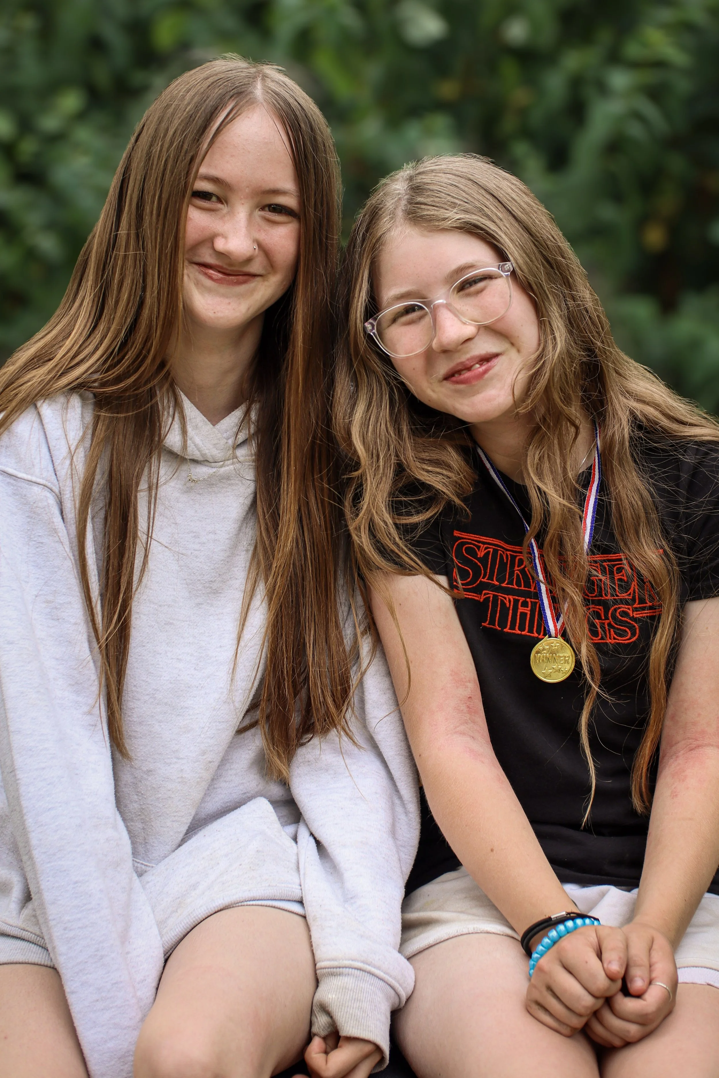 Two teenage girls sitting outdoors, smiling, with trees in the background. One girl has long brown hair, glasses, and a black 'Stranger Things' t-shirt, wearing a gold medal. The other has long straight hair, a nose piercing, and a light-colored hoodie.