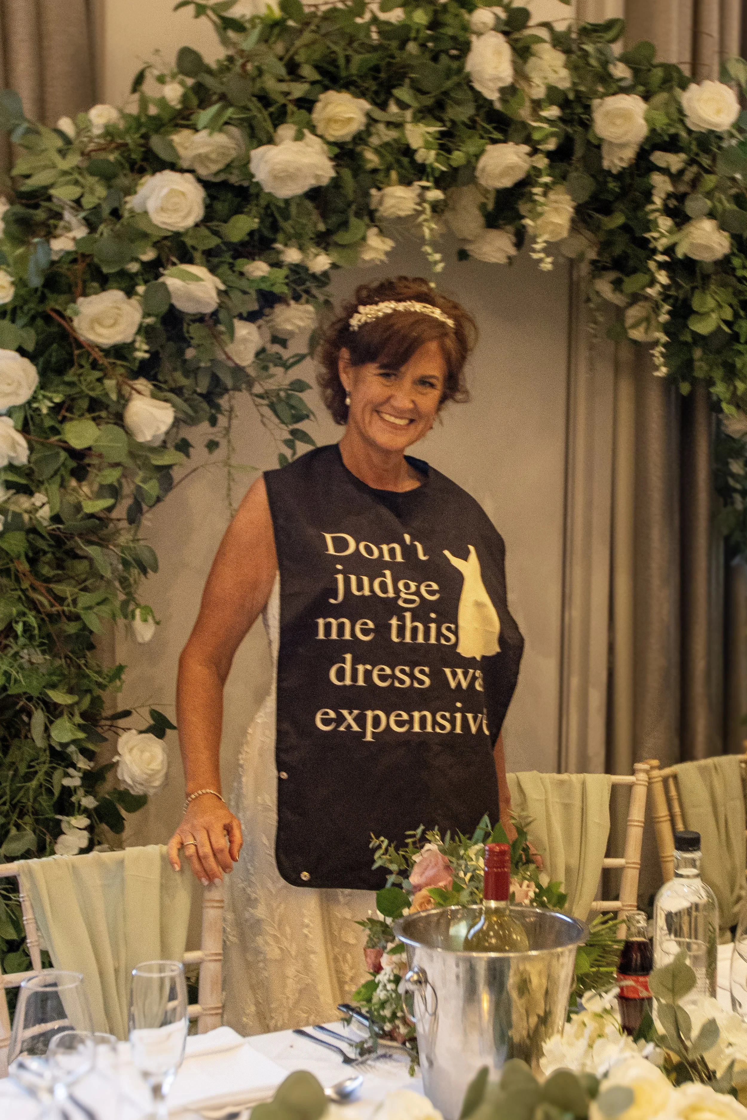 A woman smiling at a decorated table at a celebration, wearing a black apron that reads, "Don't judge me this dress was expensive," with a wallpaper of white roses and greenery in the background.