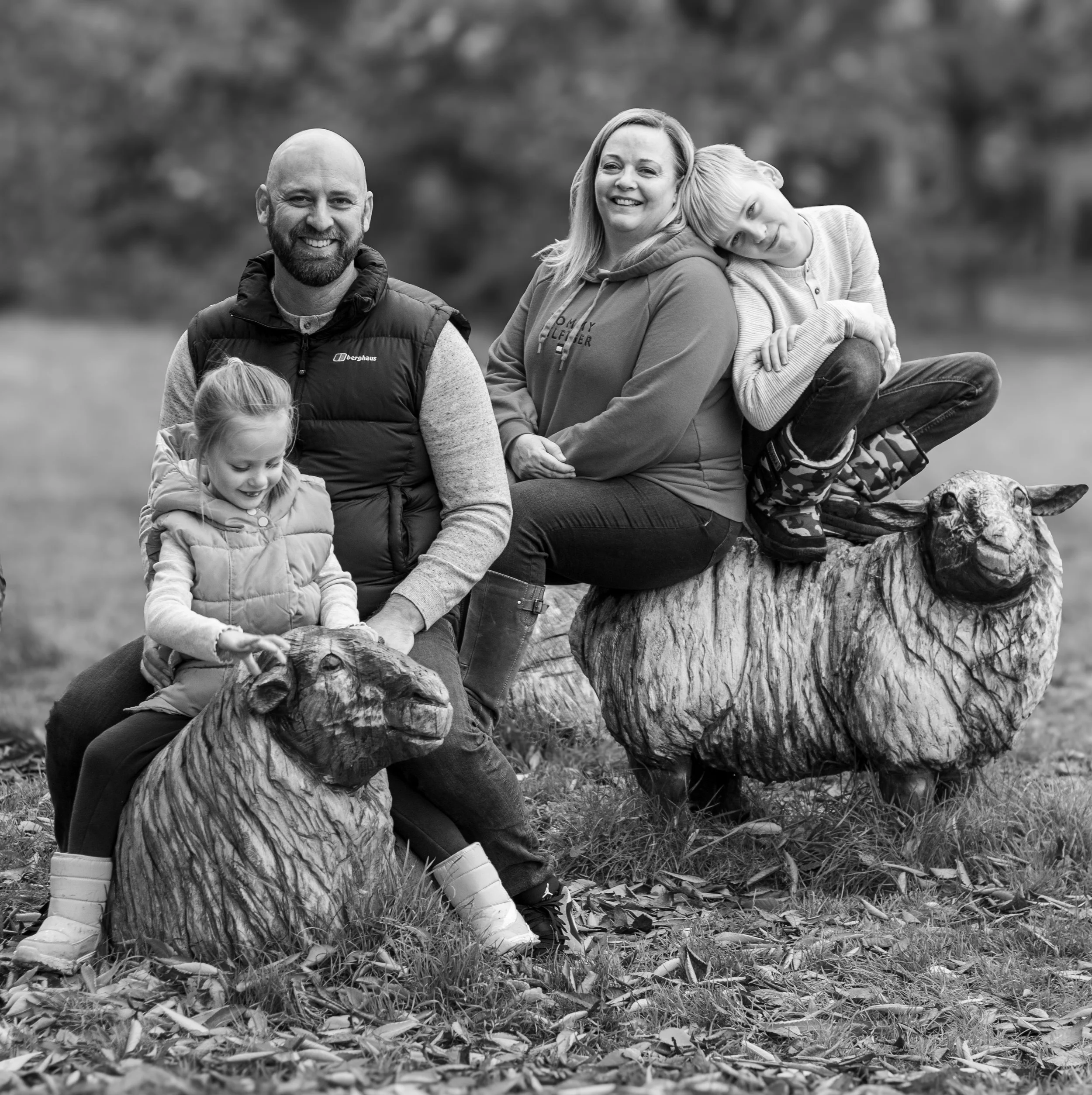 A family of four sitting on and around two wooden sheep statues outdoors, smiling and interacting with each other.