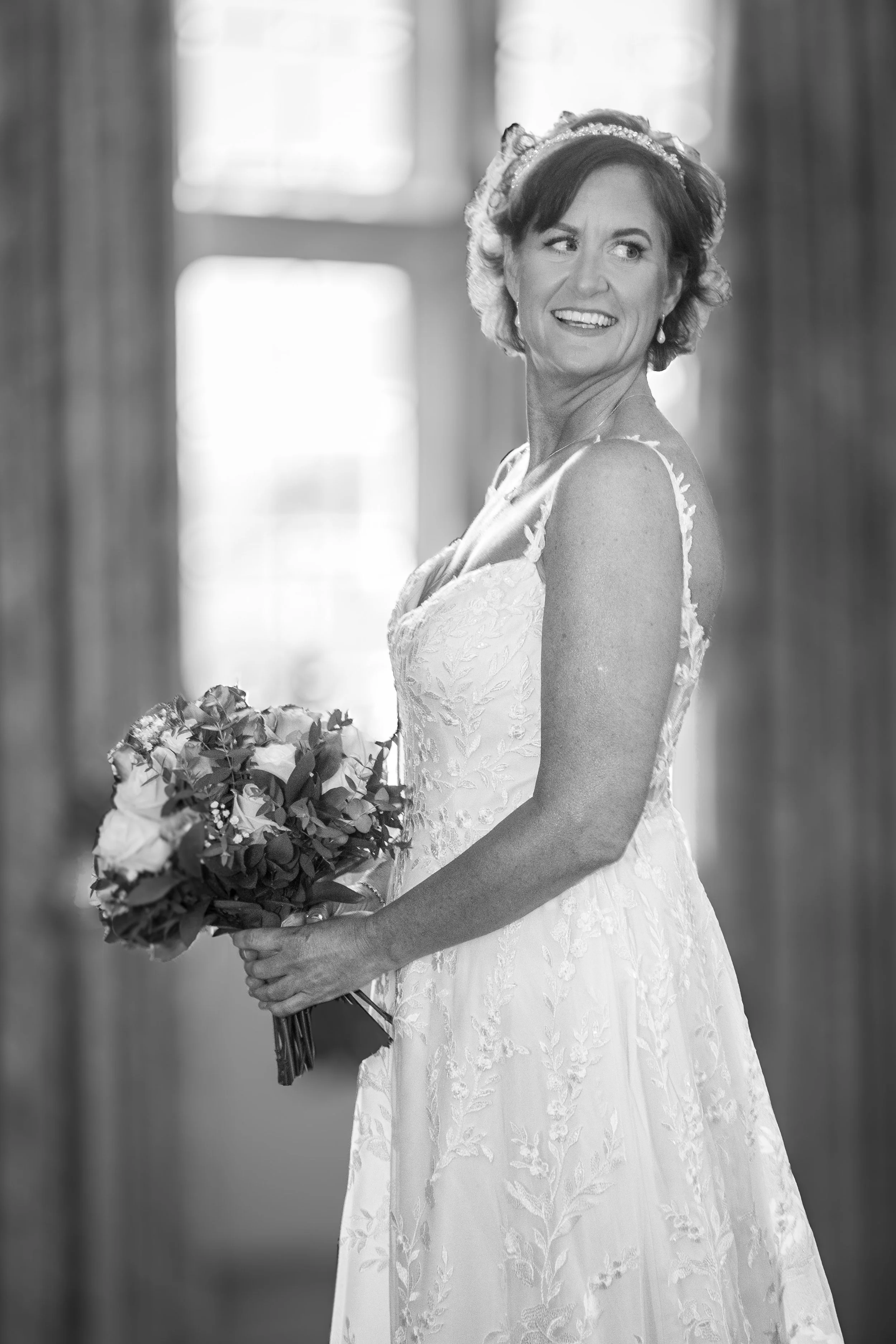 A woman in a wedding dress holding a bouquet, standing indoors with sunlight coming through windows, smiling and looking to her side.