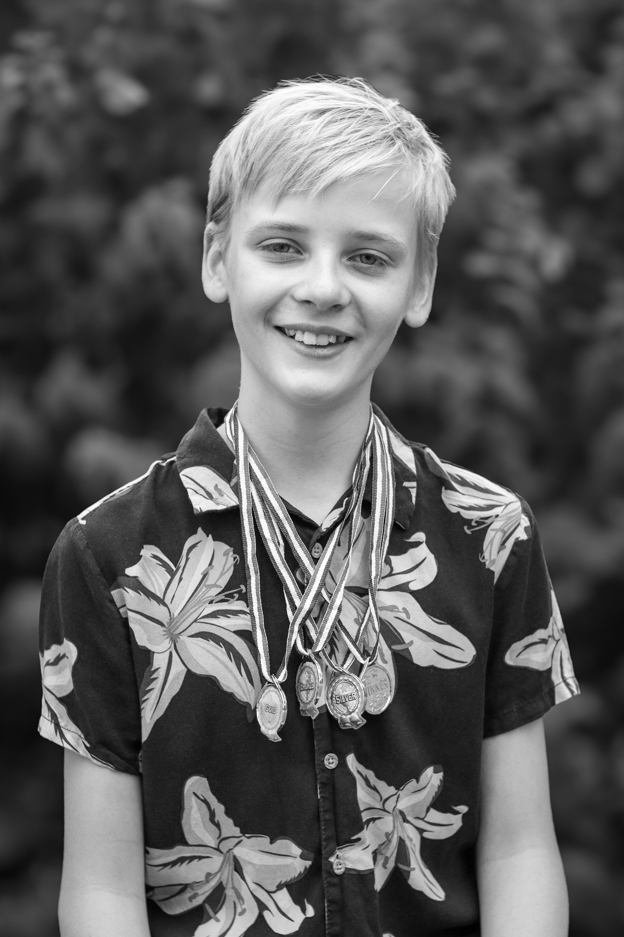 A smiling young boy with blonde hair wearing a floral shirt and multiple medals around his neck, standing outdoors with blurred foliage in the background.