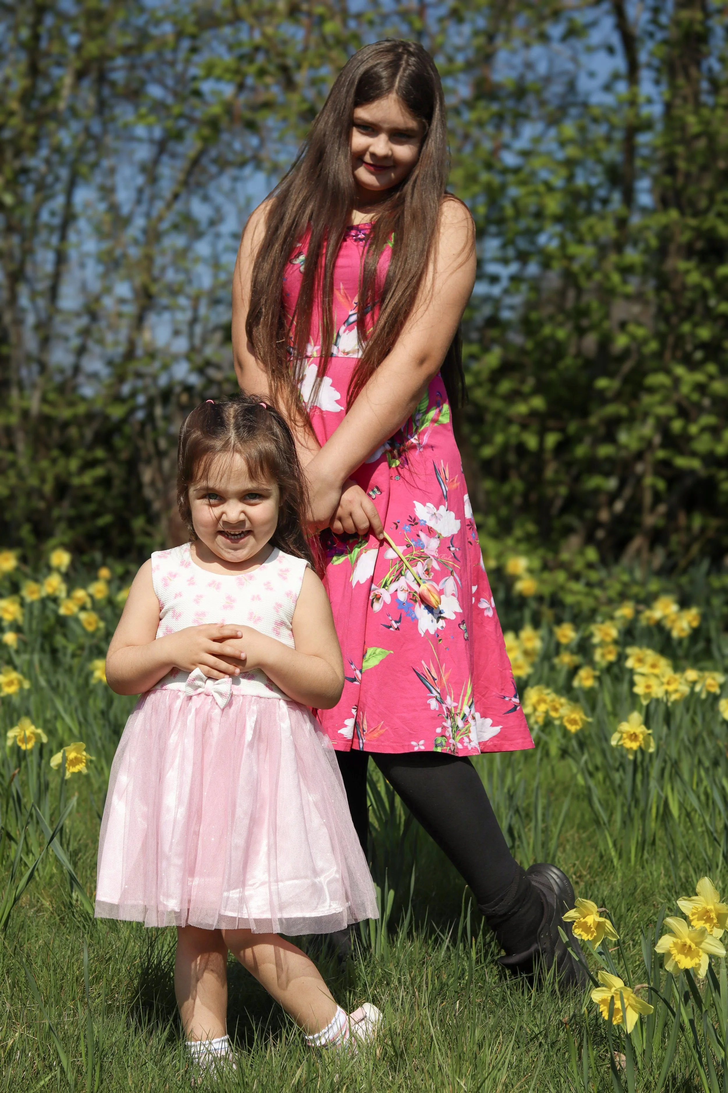Two young girls, one older with long brown hair wearing a pink floral dress, and a younger with brown hair in a white and pink dress, standing outdoors in a grassy area with yellow flowers and green trees in the background, enjoying a sunny day.