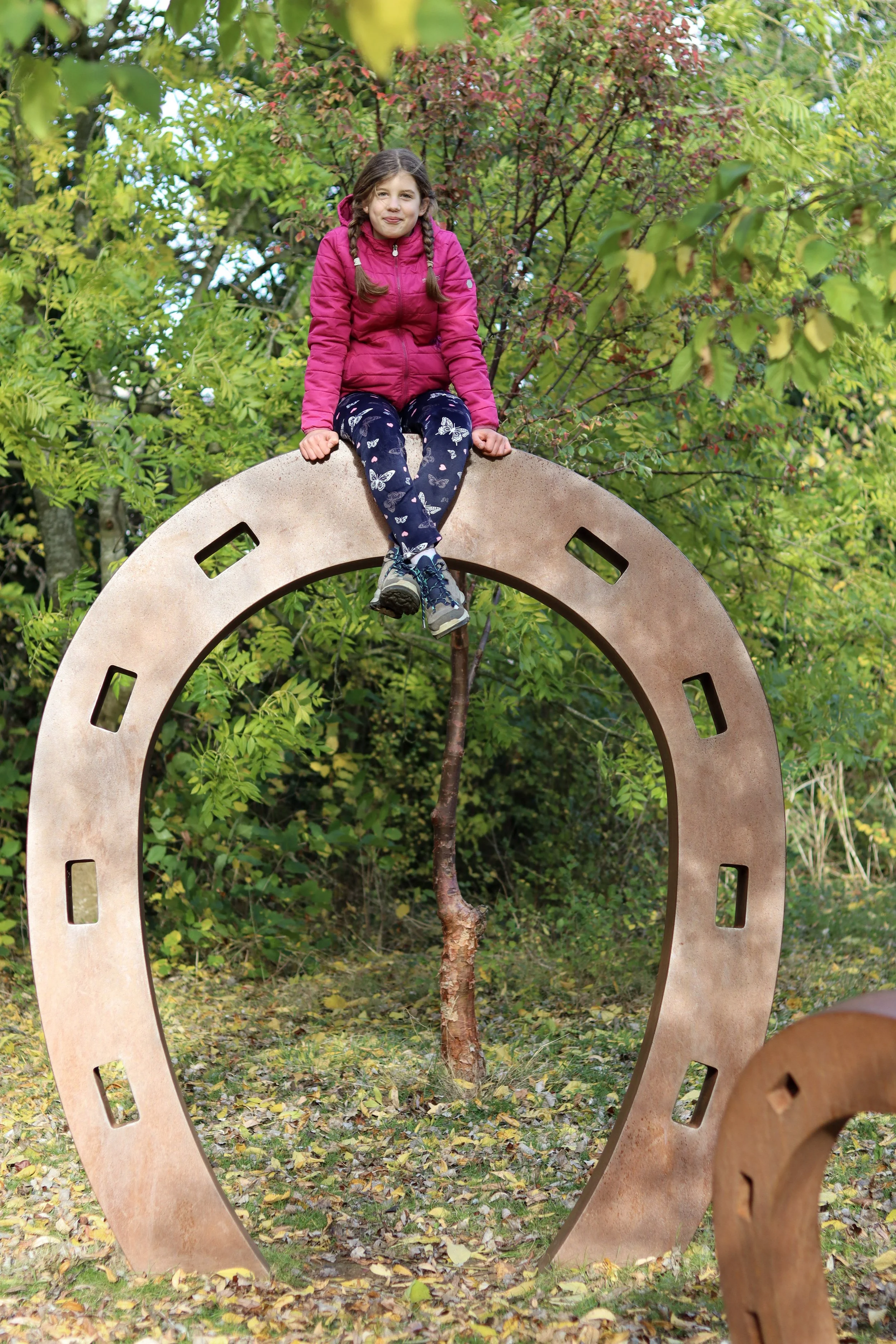 A young girl with pigtails, wearing a pink jacket and butterfly-patterned pants, sitting atop a large gear-shaped sculpture outdoors surrounded by green and yellow foliage.