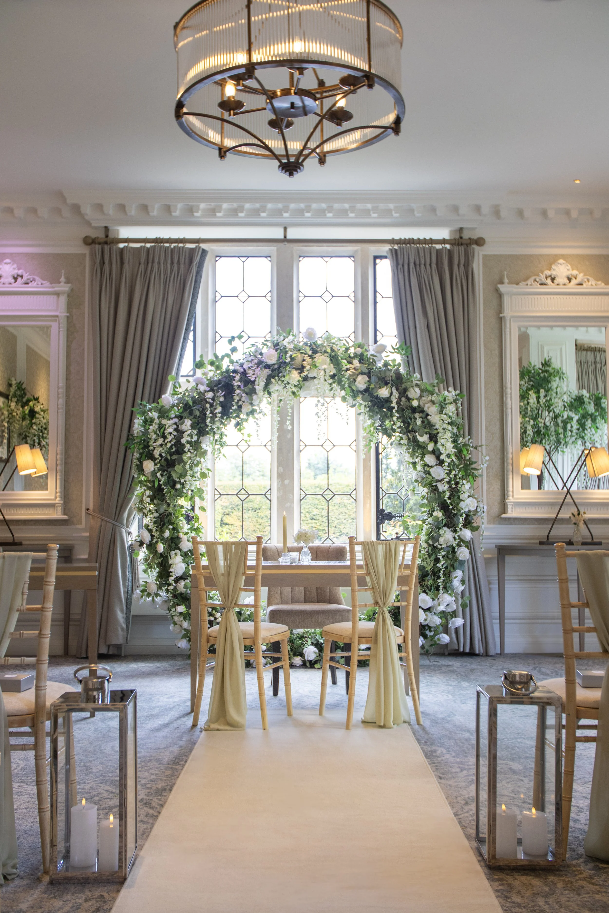 Indoor wedding ceremony setup with a floral arch, wooden chairs, a small table, and candles in glass lanterns.