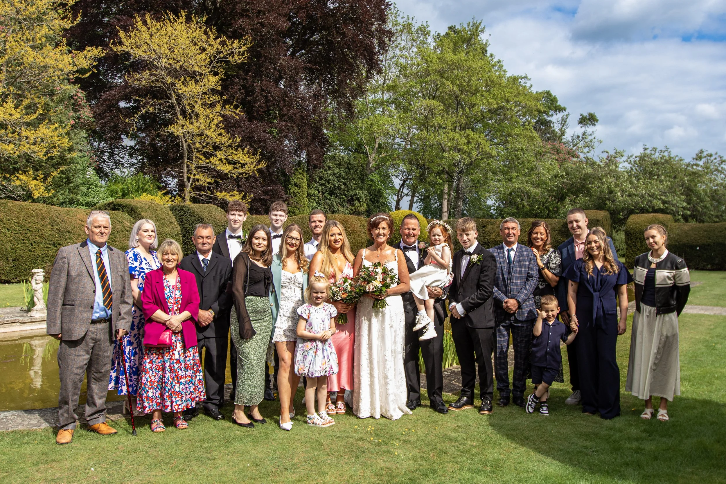 A large group of people, including a bride in a white dress holding a bouquet, posing outdoors in a garden with trees and bushes in the background during a wedding celebration.