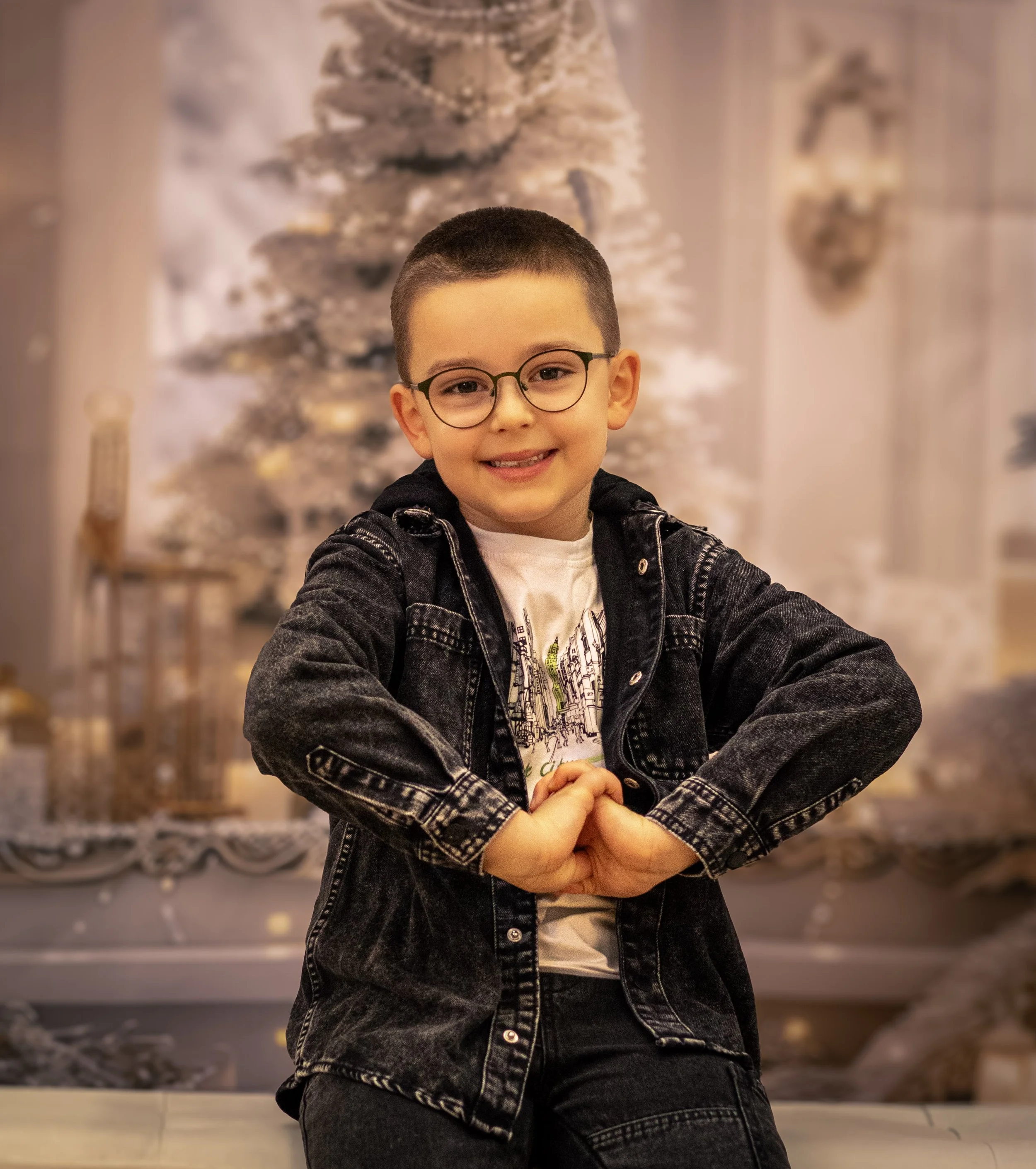 A young boy with short dark hair, glasses, and a black denim jacket smiling with hands clasped in front of a snowy Christmas tree decorated with ornaments and lights.