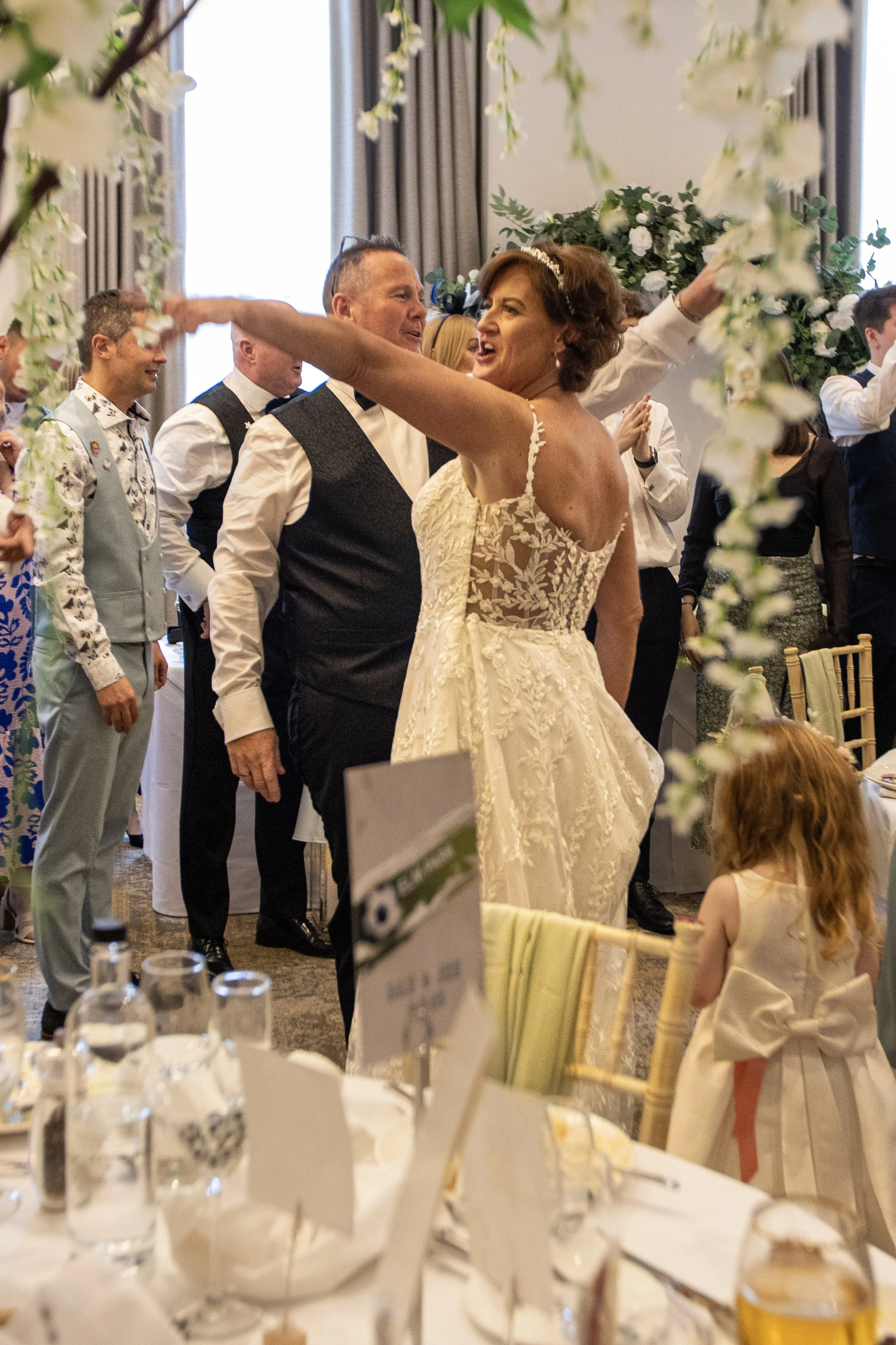 A bride and groom dancing at a wedding reception surrounded by guests, with a young girl with red hair watching nearby.