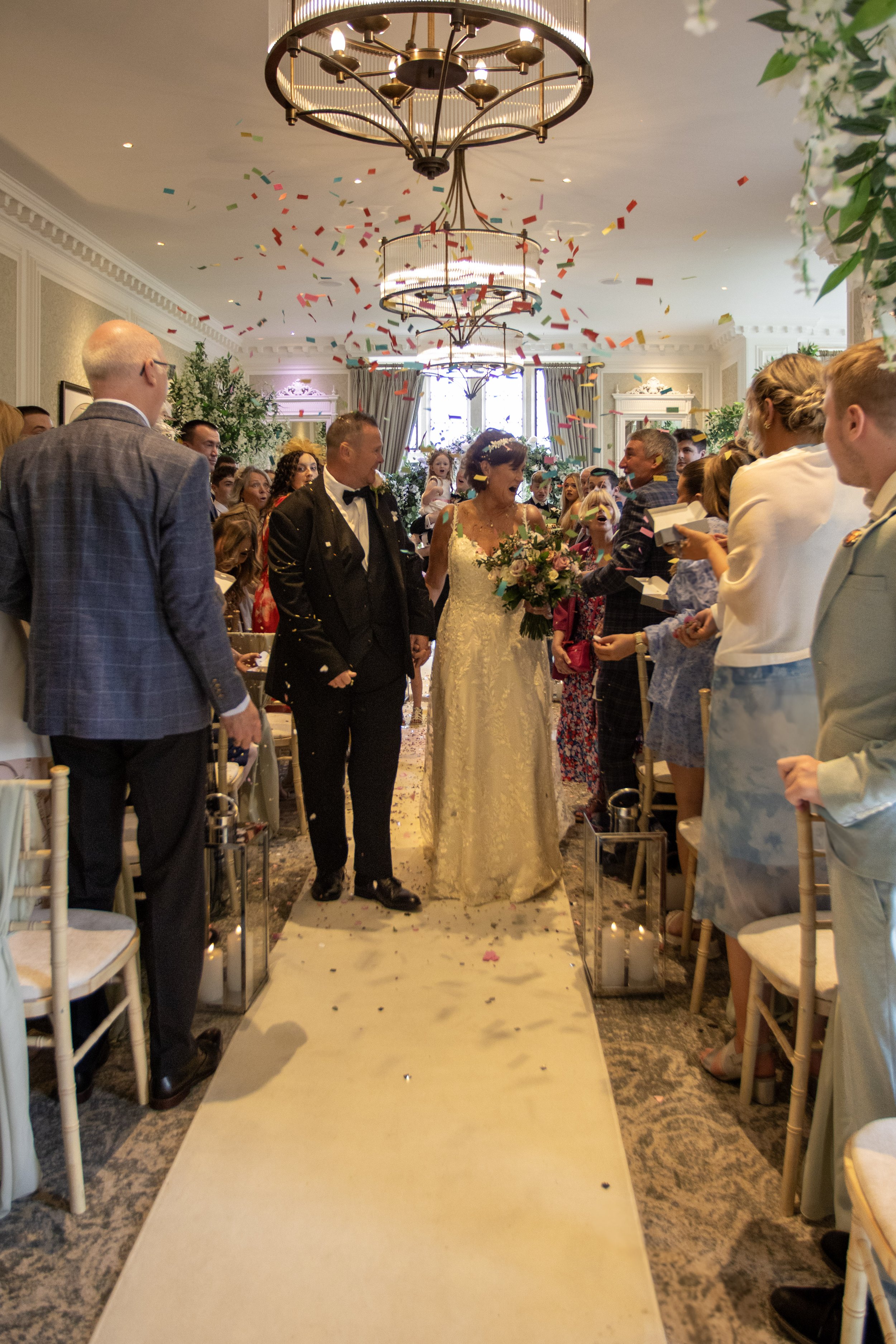 Bride and groom walking down the aisle at a wedding ceremony, surrounded by guests. Confetti is falling from the ceiling, and there are chandeliers above.
