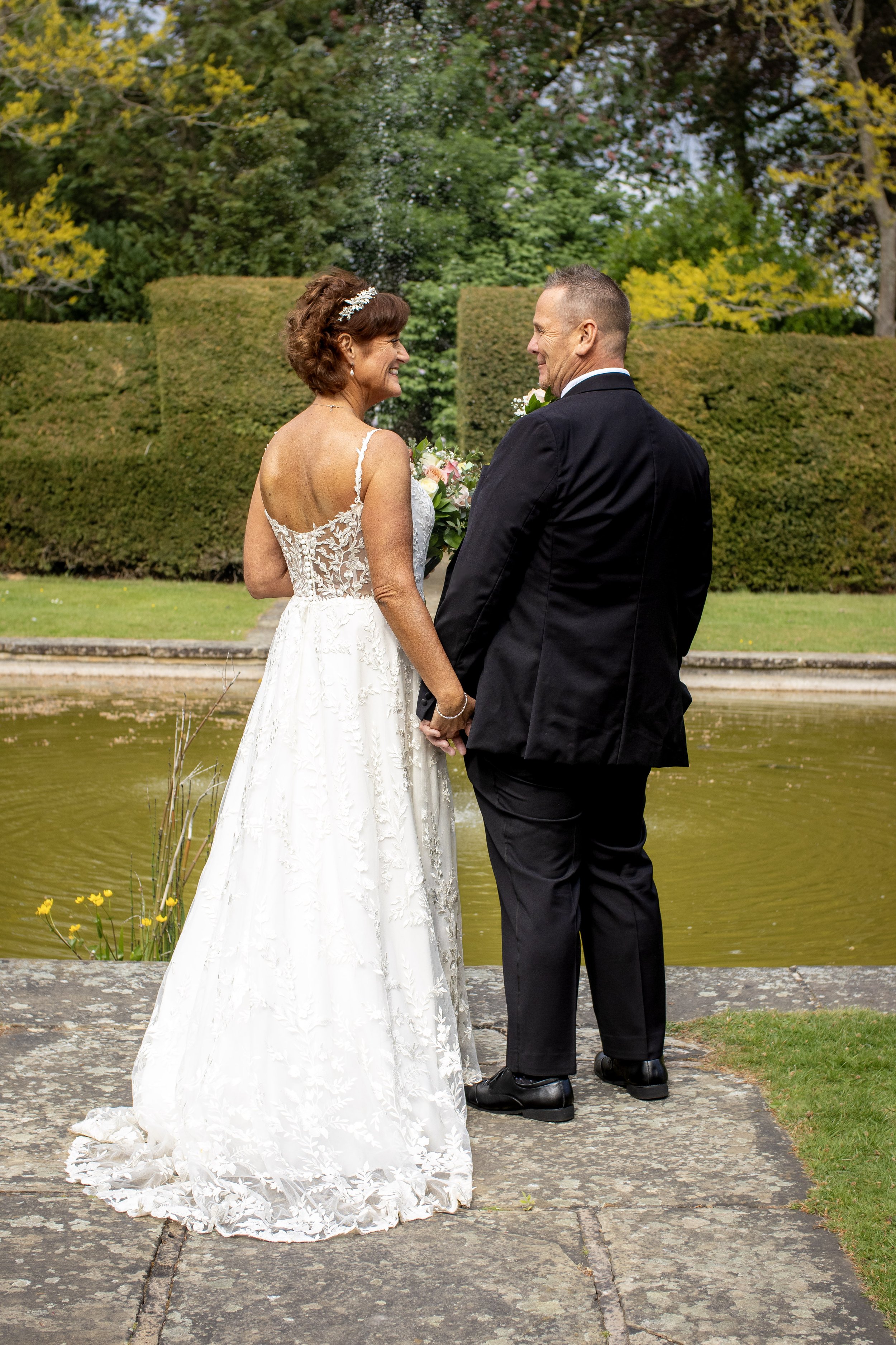 A bride and groom holding hands and smiling at each other outdoors near a pond with lush green trees and bushes in the background, on a cloudy day.