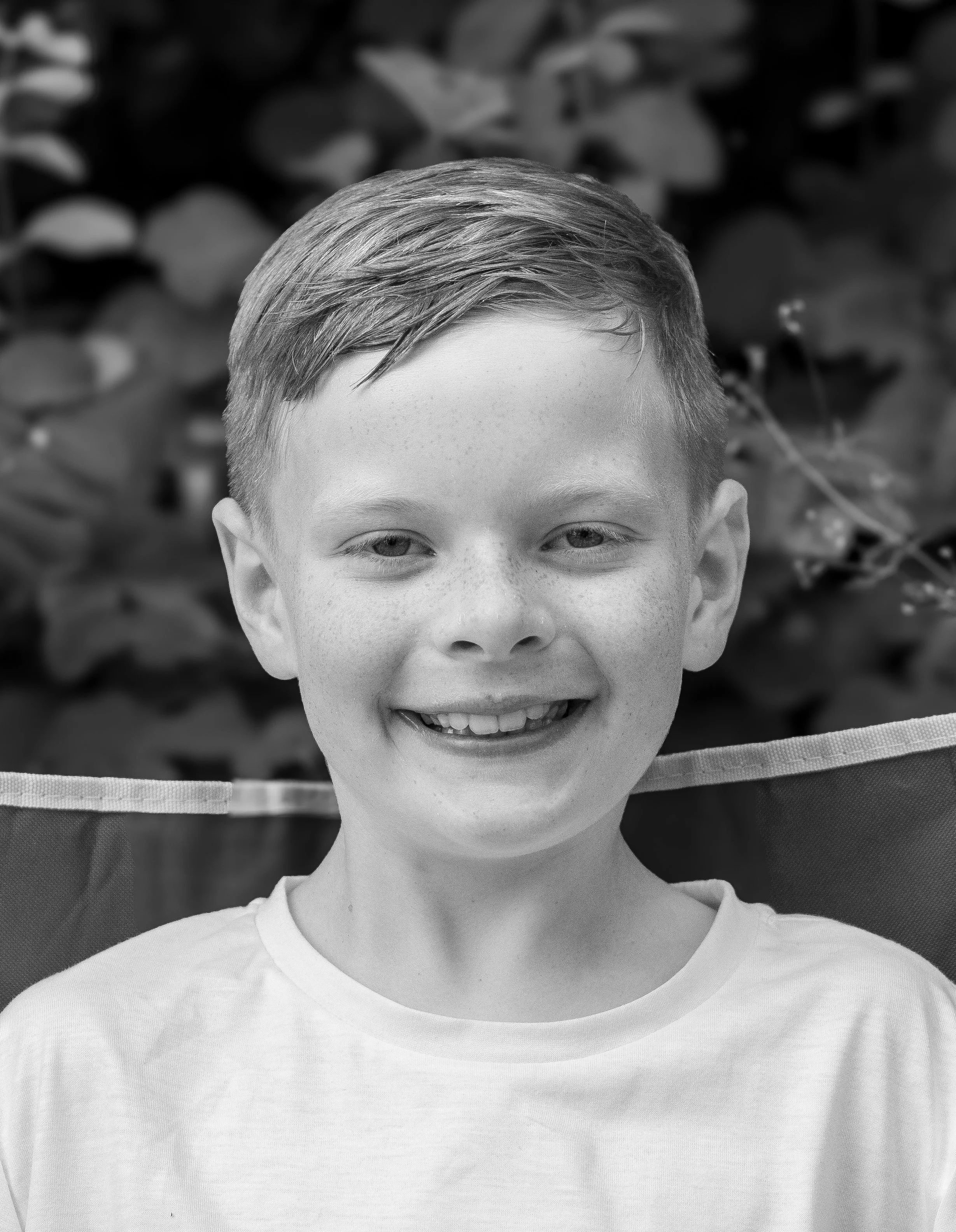 Smiling young boy with short hair and freckles, sitting outdoors with blurred foliage in the background.
