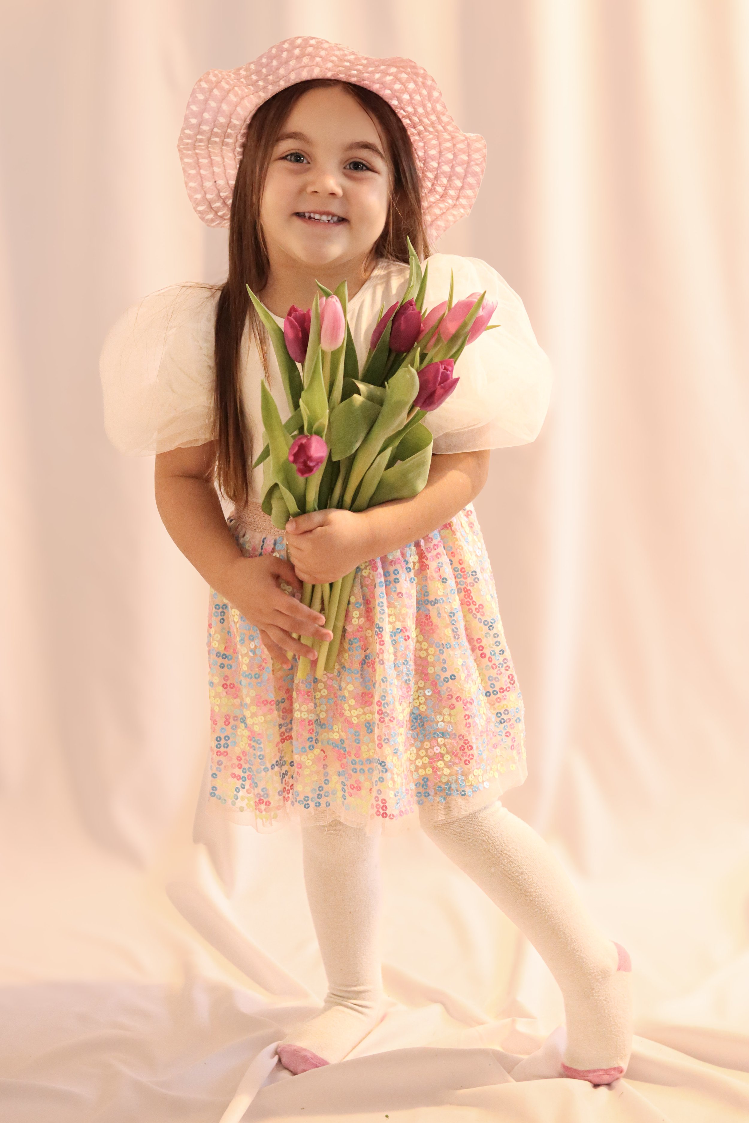 A young girl wearing a pink sun hat, a pastel dress with puffed sleeves, and white tights, holding a bouquet of pink tulips, smiling at the camera with a light-colored backdrop.