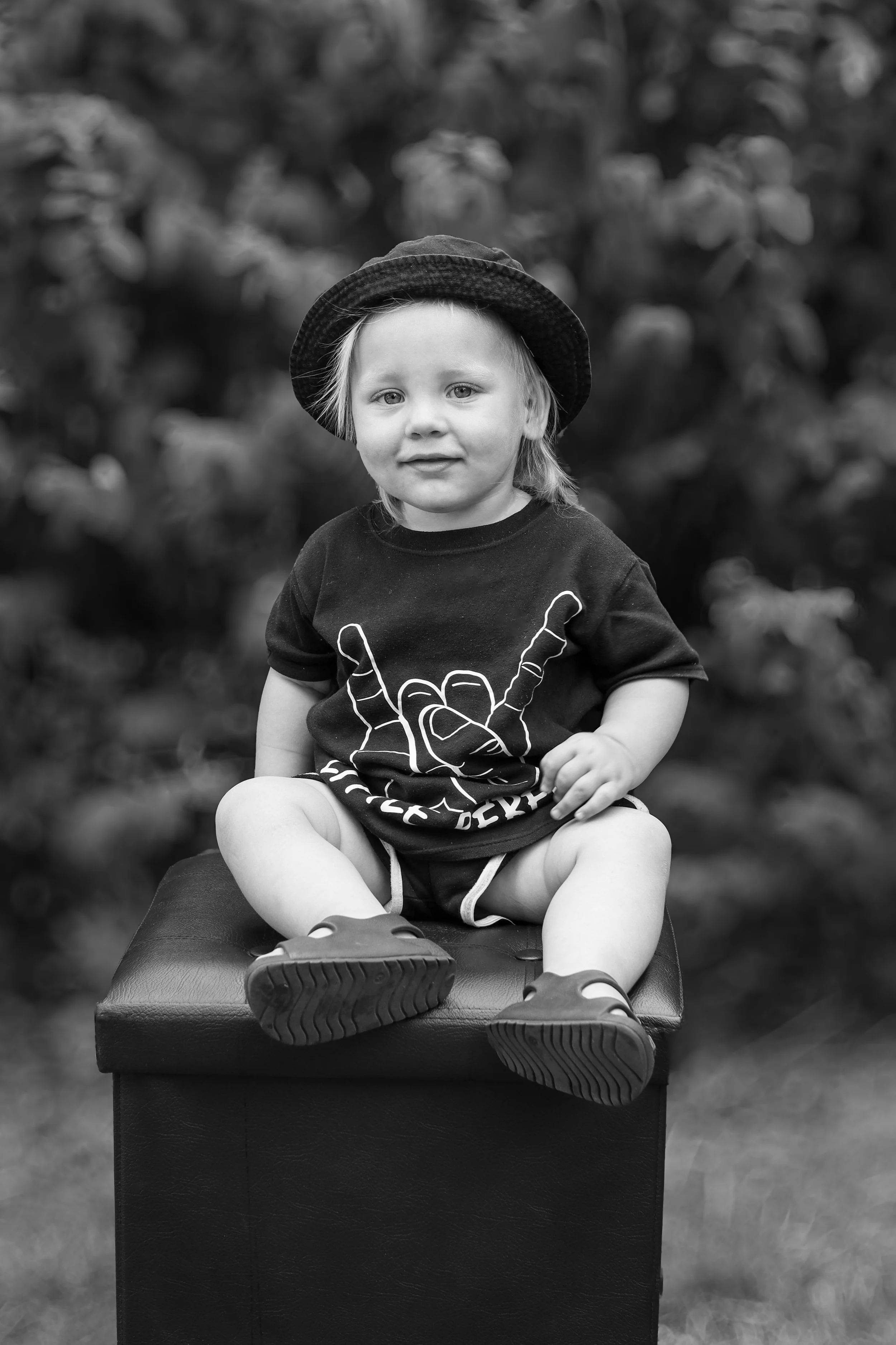 A young child with light hair, wearing a hat and a T-shirt with a hand making a peace sign, sitting on a box outdoors, looking at the camera, in black and white.