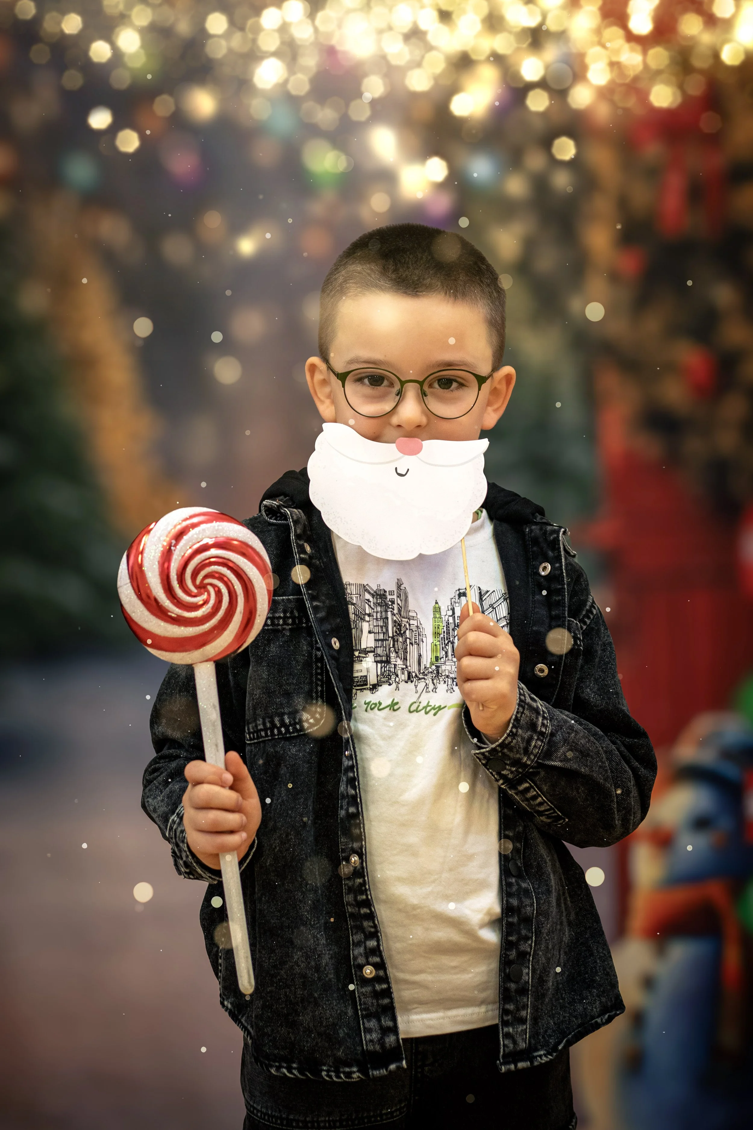 A young boy with glasses holds a red and white peppermint lollipop and a paper cutout of Santa's face in front of a blurred festive background.