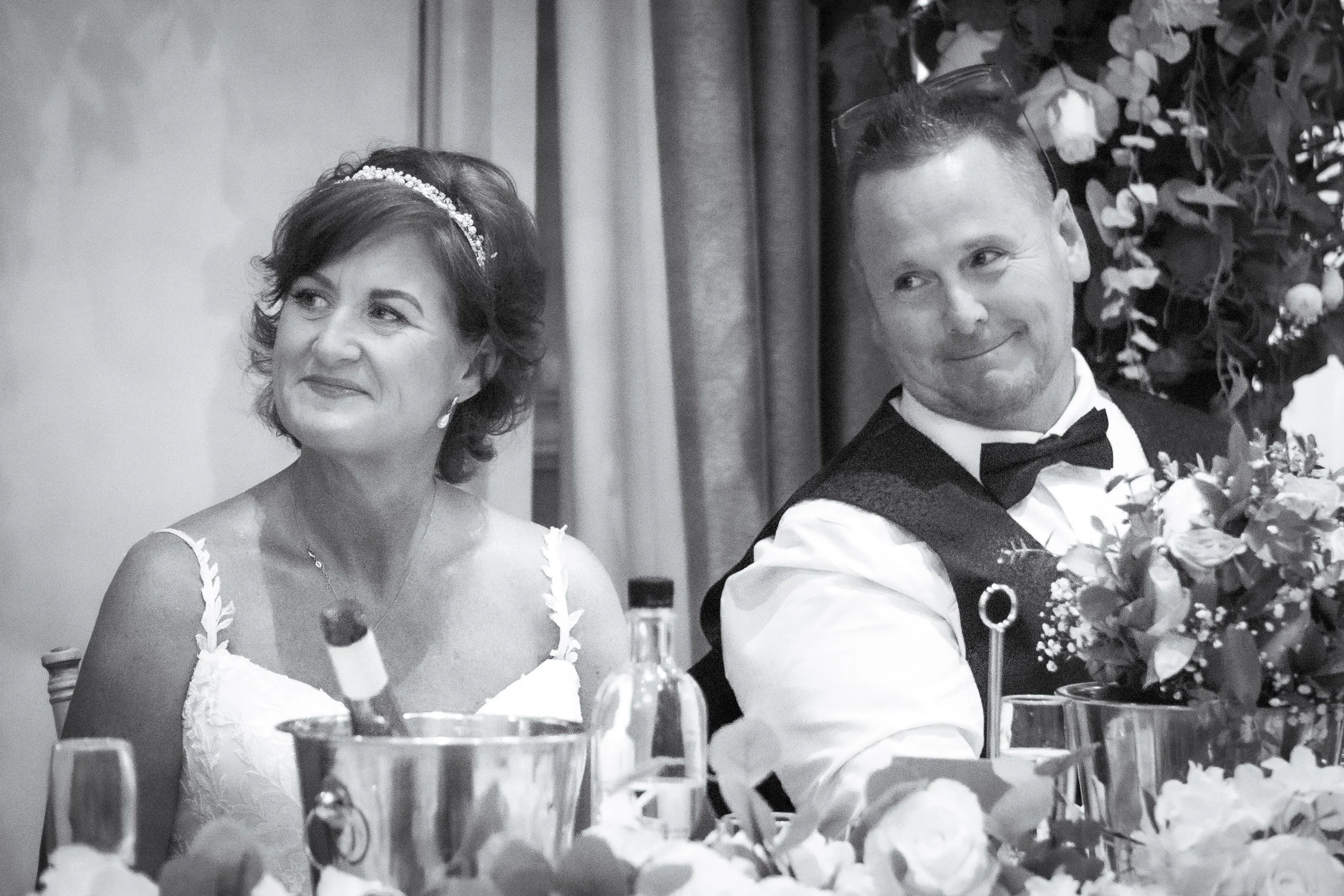 A woman in a wedding dress and a man in formal attire sitting at a table, smiling and looking in different directions, surrounded by flowers and bottles.