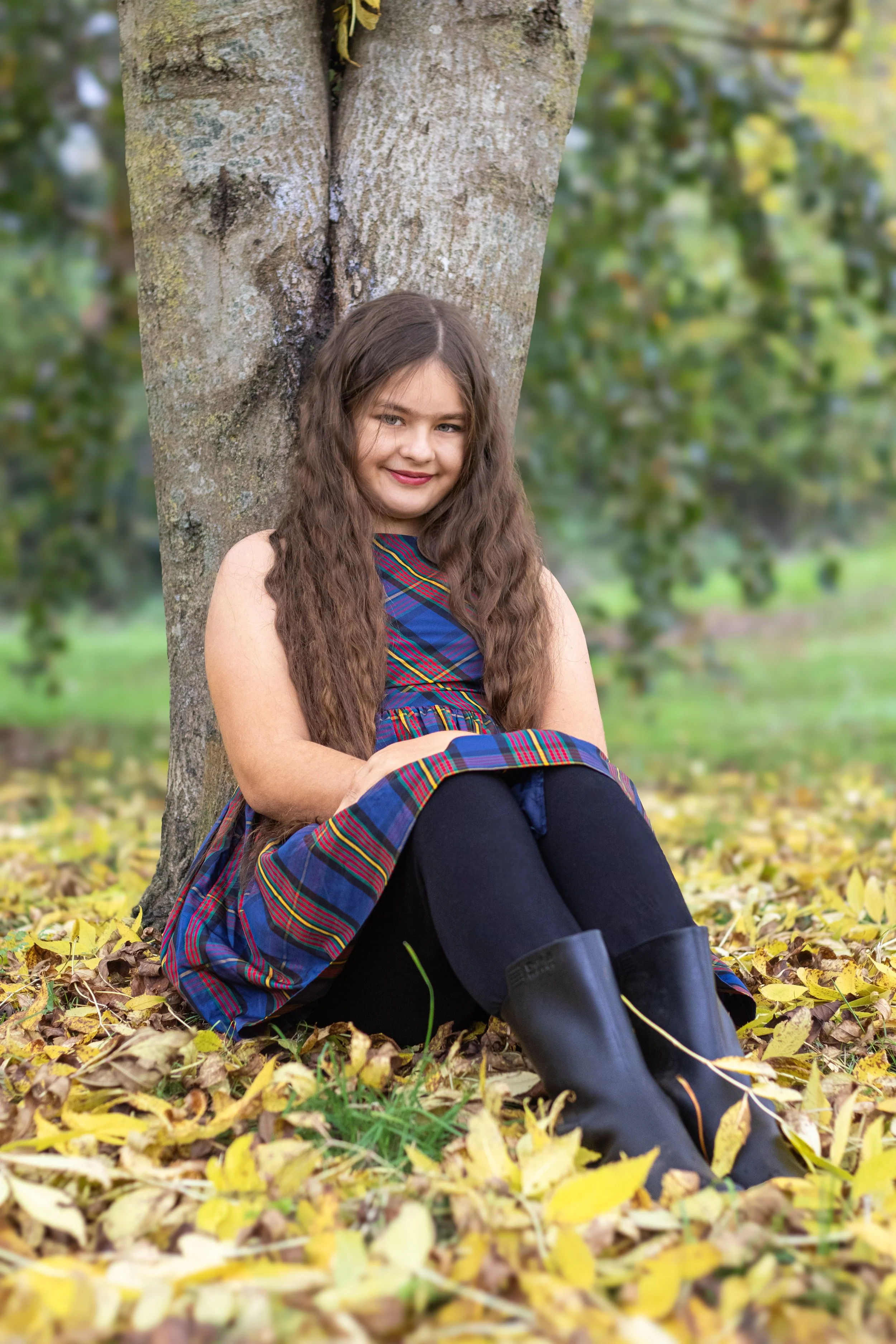A young girl with long, wavy brown hair sitting against a tree in a park during fall, surrounded by yellow leaves, wearing a plaid dress, black leggings, and black boots, smiling at the camera.