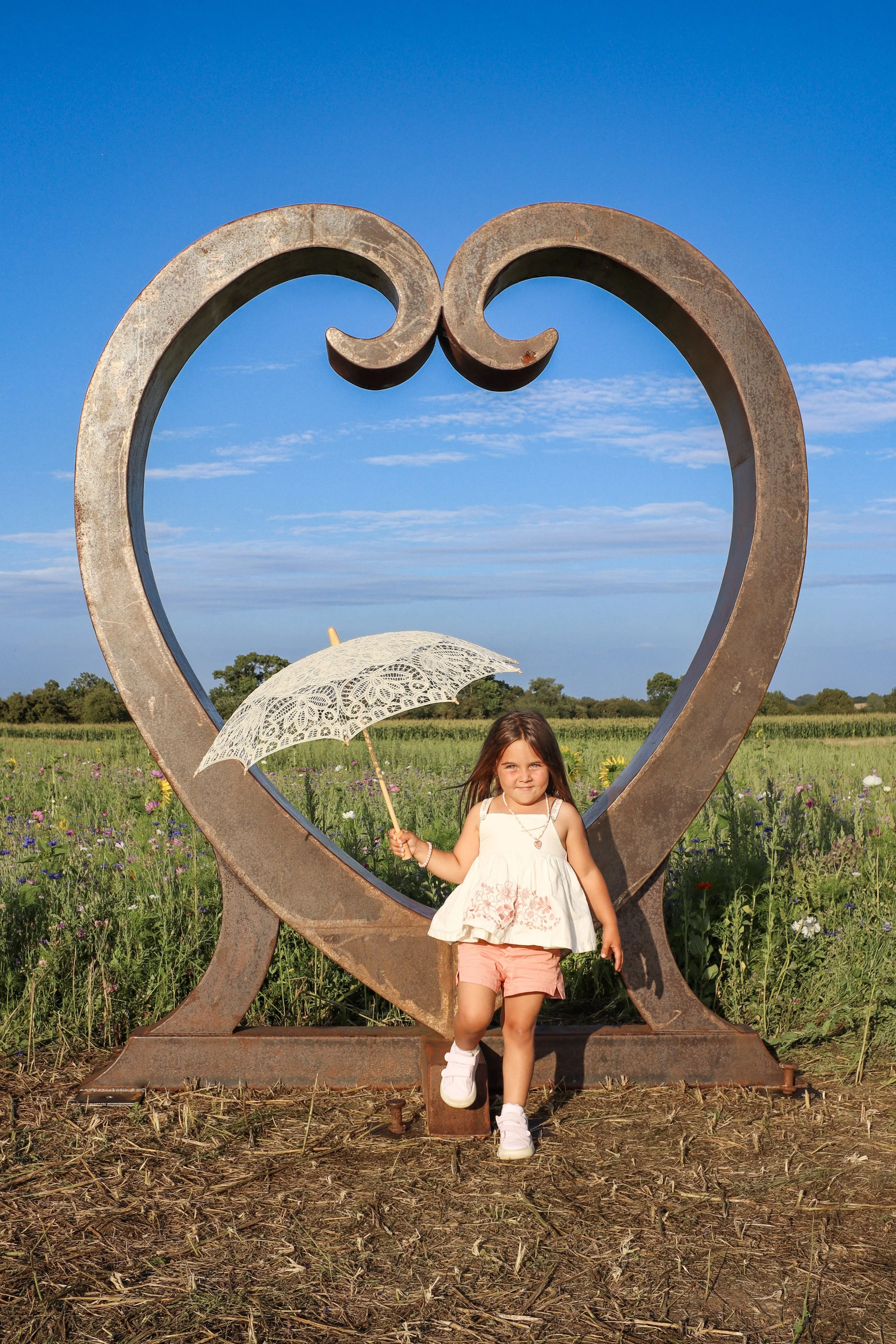 A young girl in a white dress and pink shorts holding a lace umbrella is standing inside a large metal heart-shaped sculpture in a field of wildflowers under a bright blue sky.