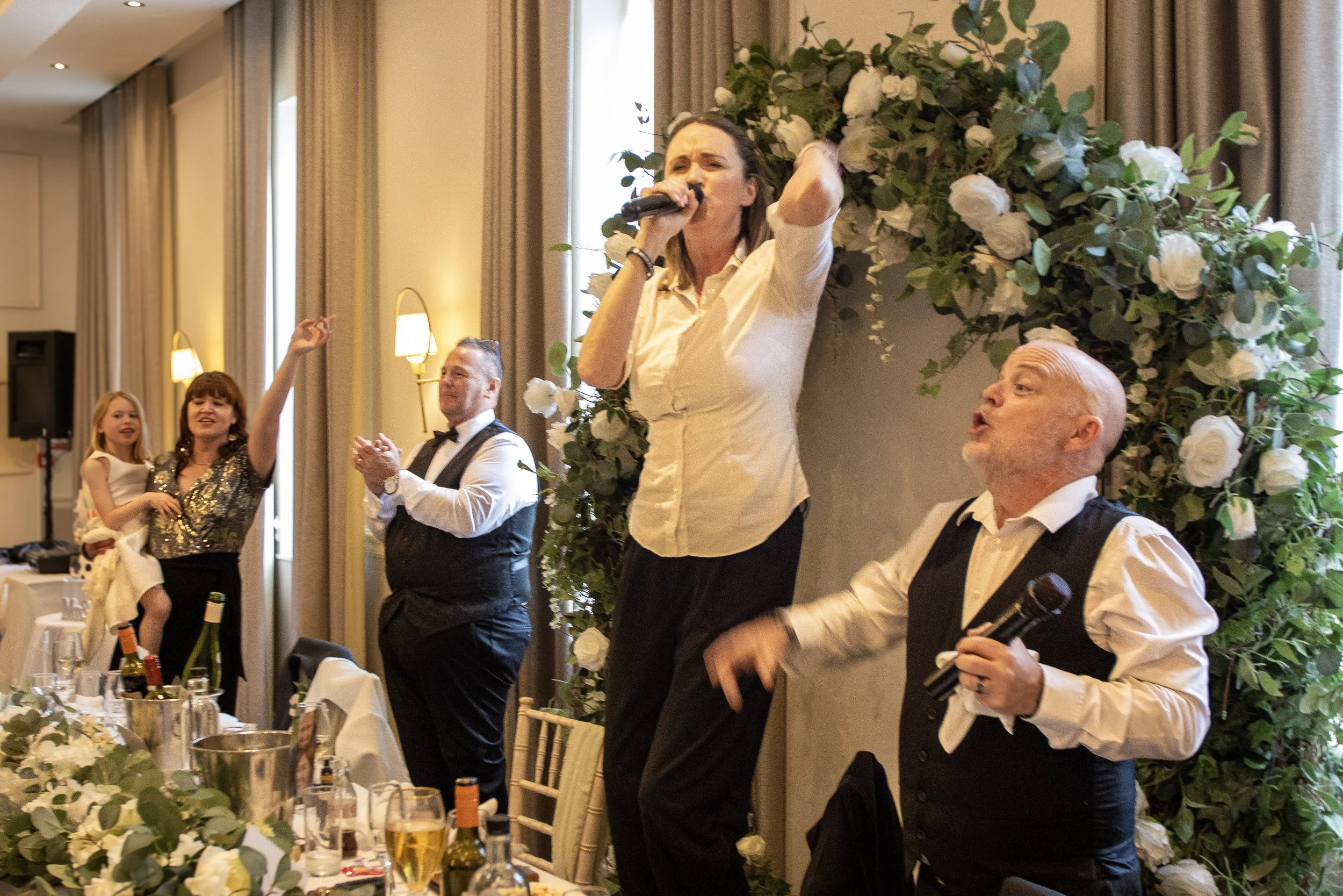 People celebrating at a wedding reception, with a woman singing into a microphone and a man next to her holding another microphone, in front of a floral backdrop with white roses and greenery.