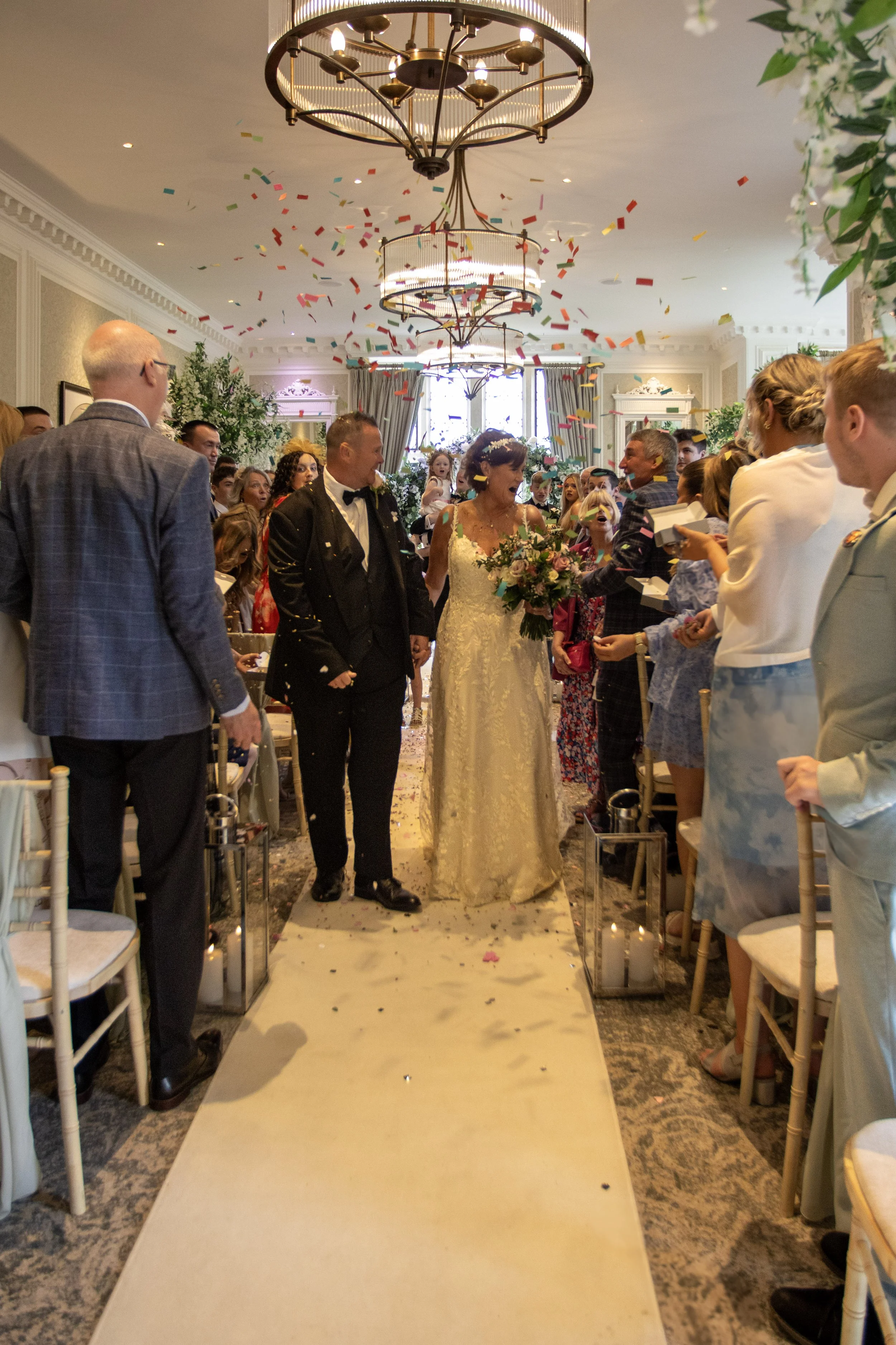 A bride and groom walking down the aisle at their wedding, surrounded by guests throwing confetti, in a decorated indoor venue with chandeliers and floral decor.
